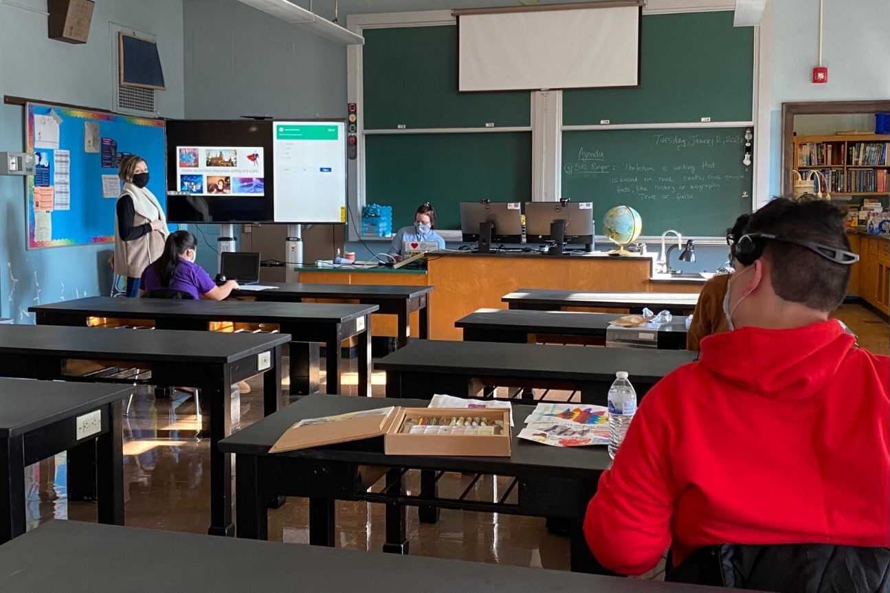 A teacher and students several feet apart sit at desks, working in a classroom