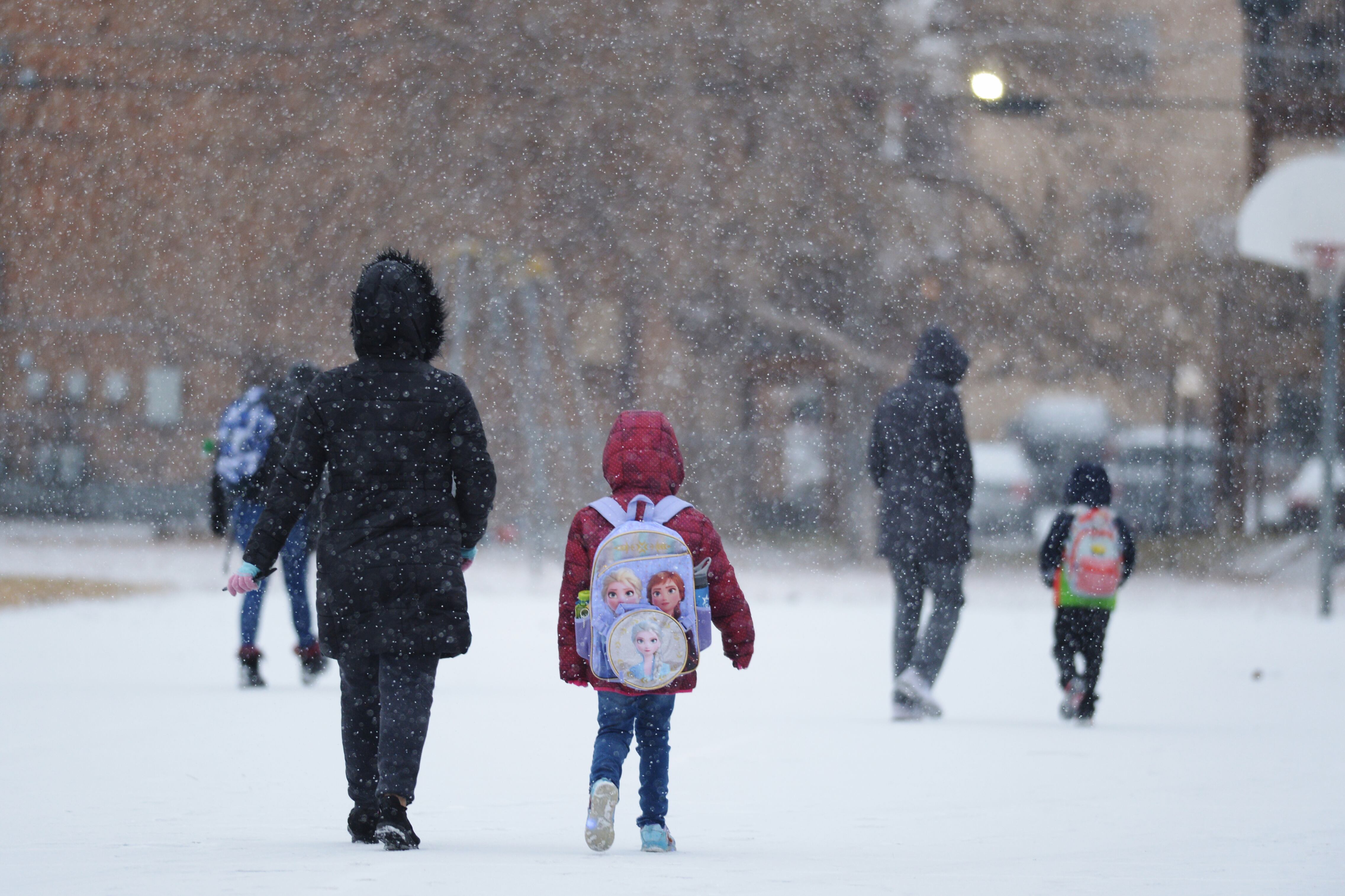Children wearing winter coats and backpacks walk to school on a snowy day.