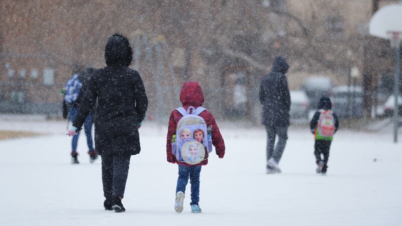 Children wearing winter coats and backpacks walk to school on a snowy day.