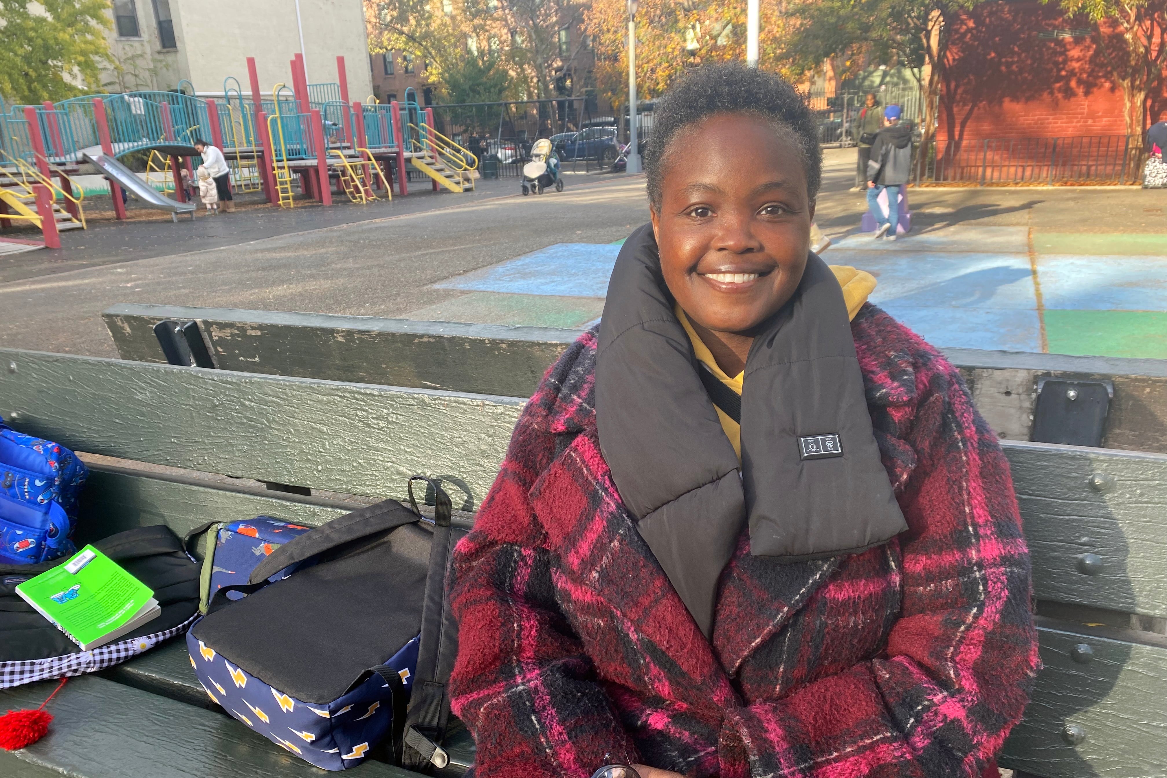 A woman with short black hair, wearing a grey scarf and a red and black jacket smiles while posing for a portrait with a playground in the background.