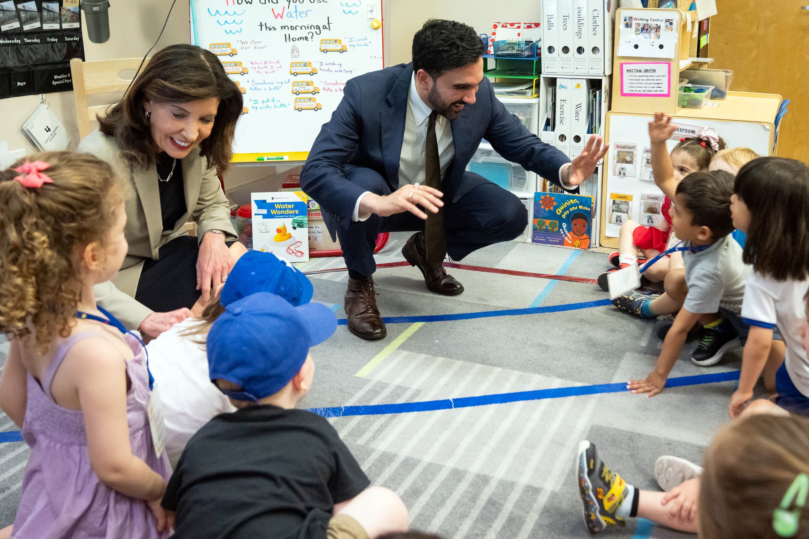 A photograph of two adults in suits kneeling down to greet young children in a colorful classroom.