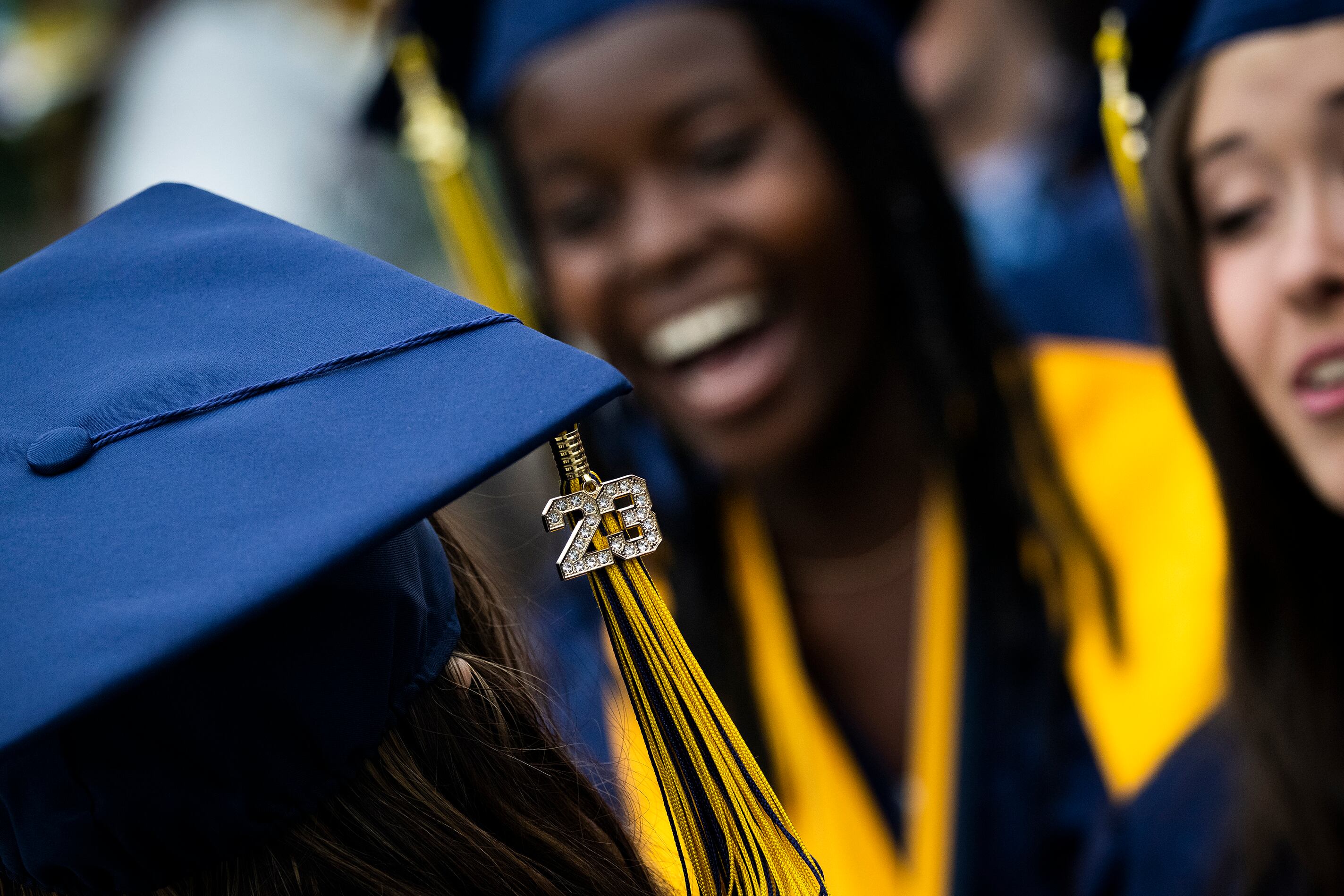 A close up of a high school graduation cap with the number 23 hanging with a tassel and two high school graduates standing in the background.
