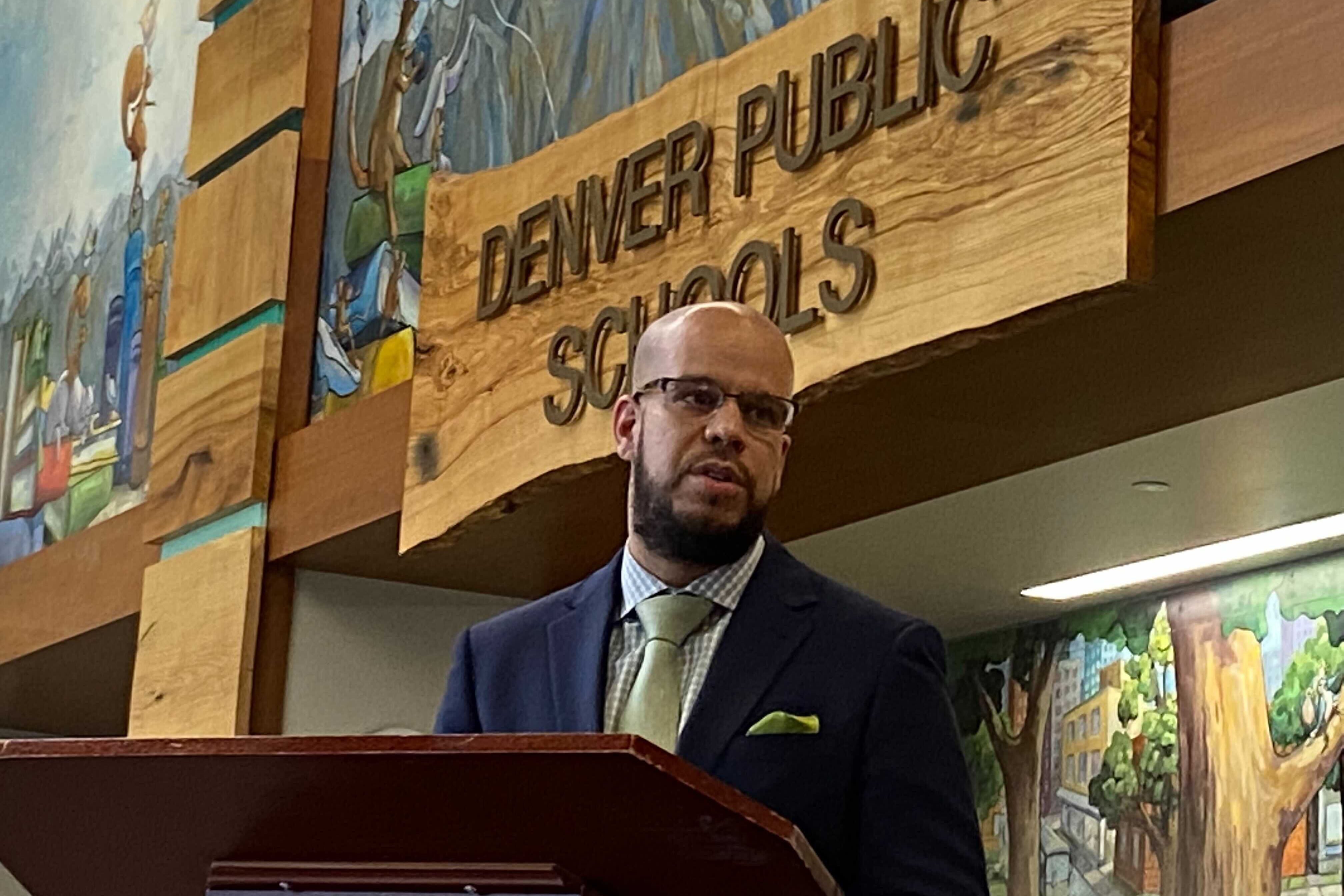 A man wearing a suit and tie stands at a podium with a sign in the background that reads, "Denver Public Schools."