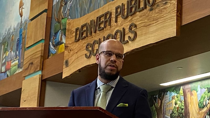 A man wearing a suit and tie stands at a podium with a sign in the background that reads, "Denver Public Schools."