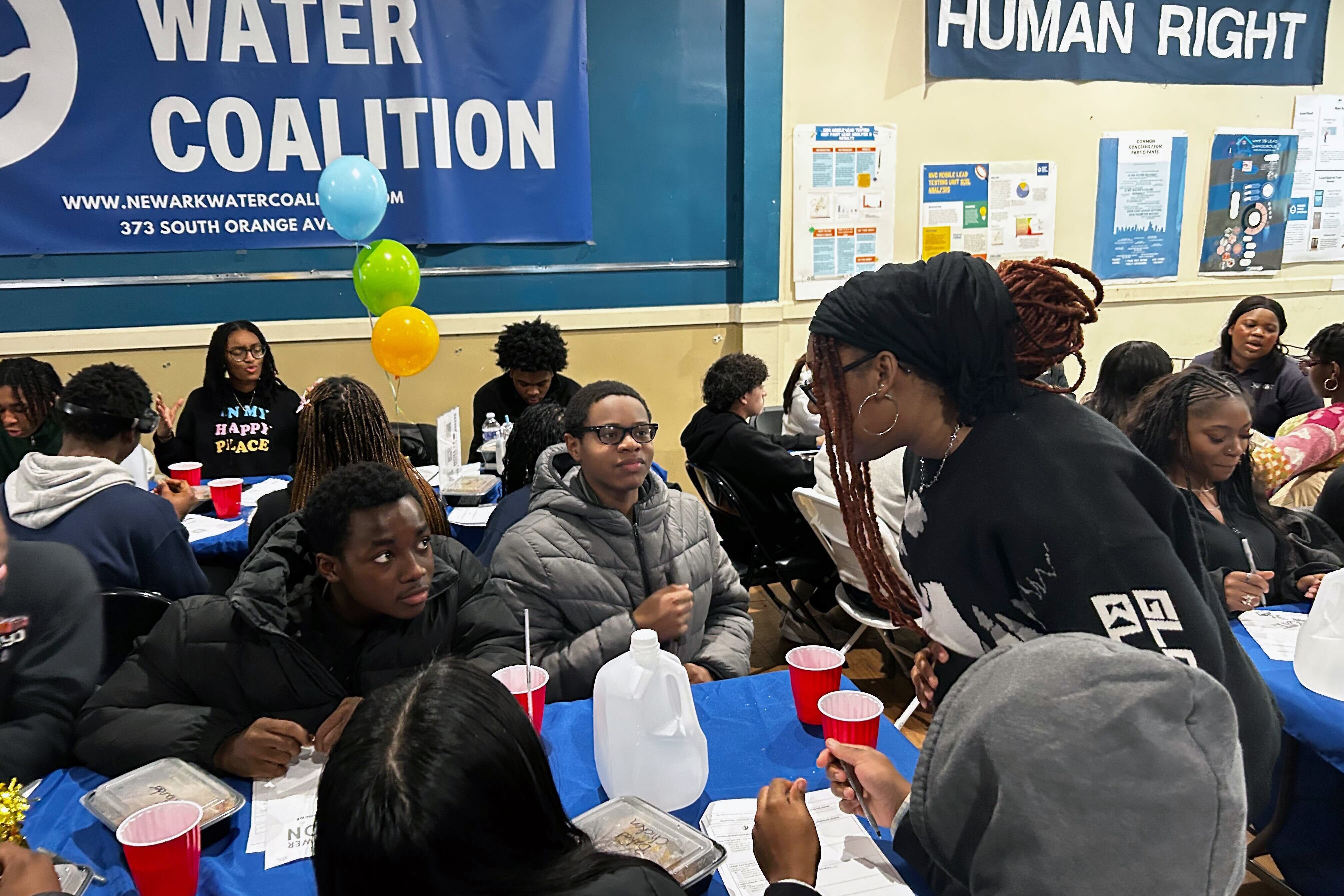 A group of high school students sit at tables with blue table cloths in a room.
