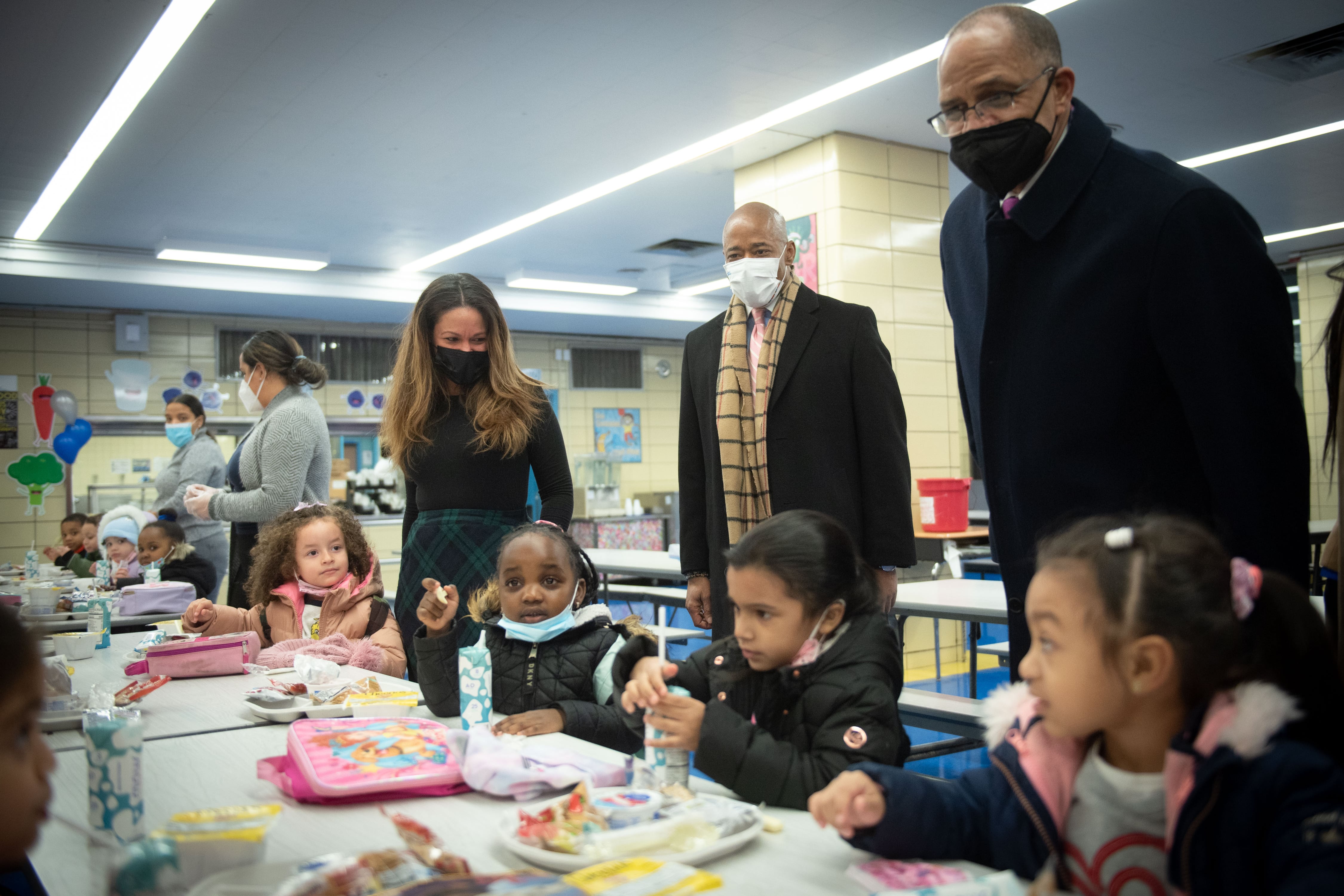 Children sit at a long table with three adults standing behind them.