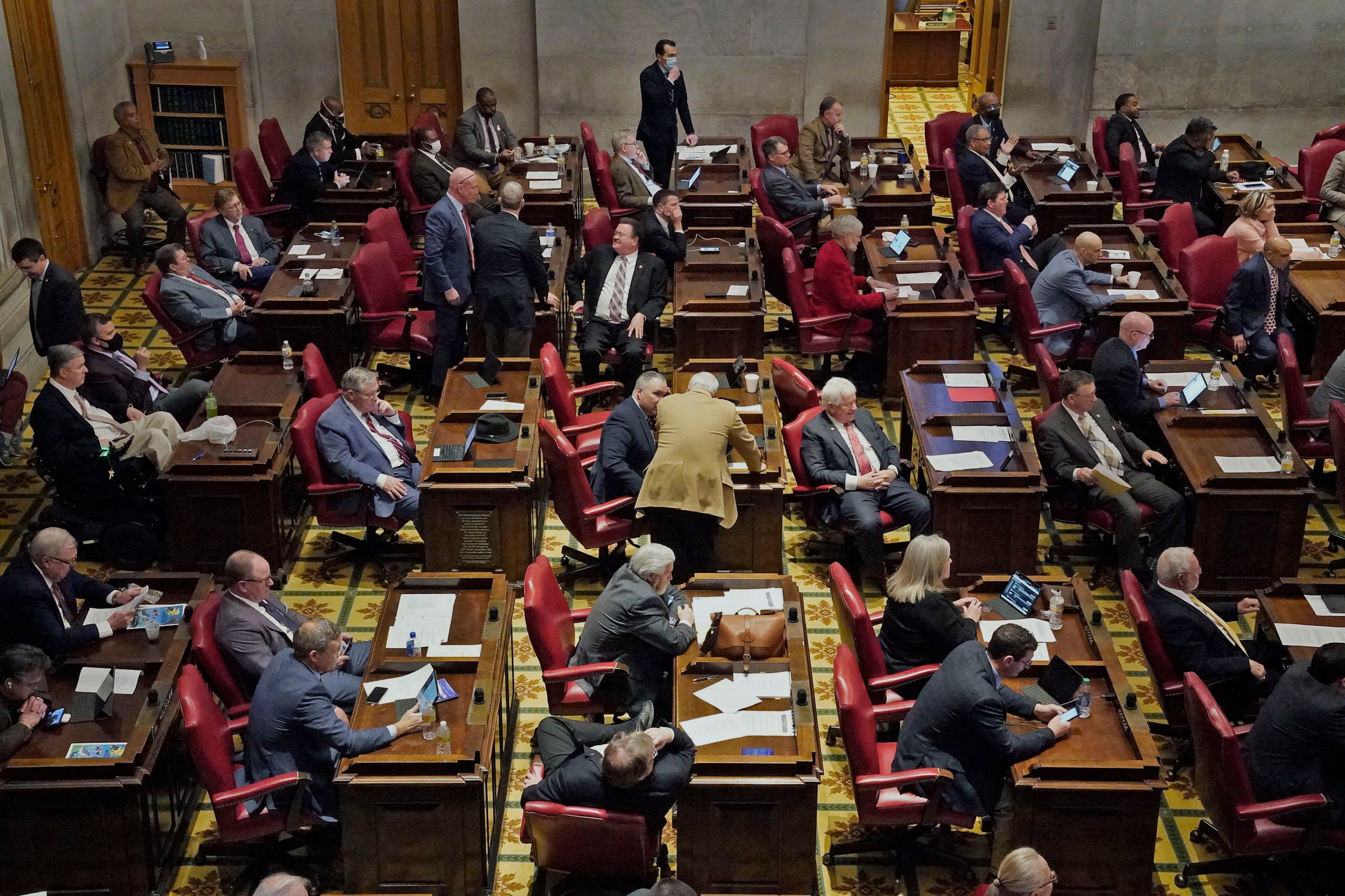 Men and women wearing suits sit behind desks in a large legislative chamber.