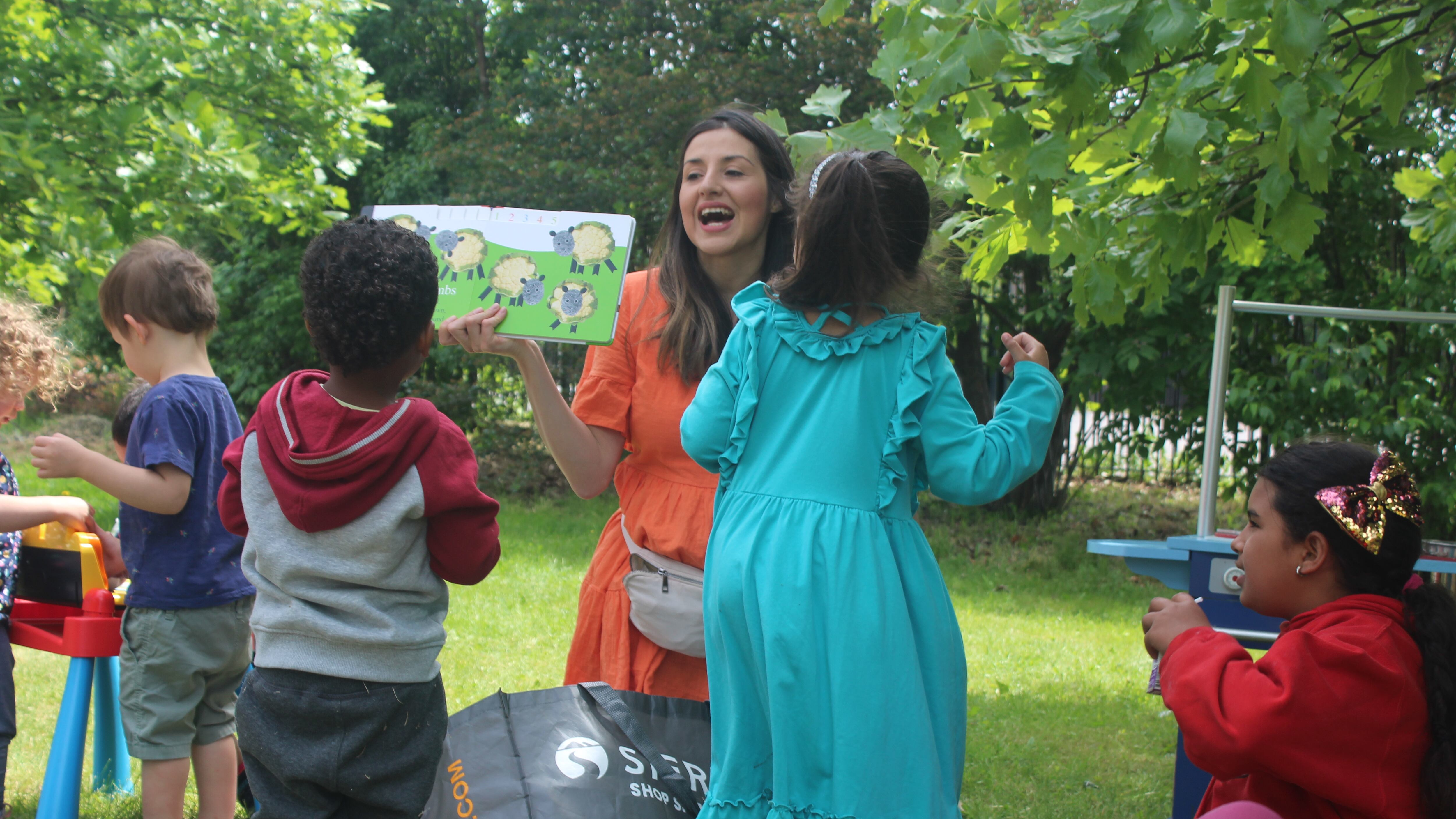 A woman with brown hair and a red dress laughs with four children of different ages while standing on grass in front of trees in a park.