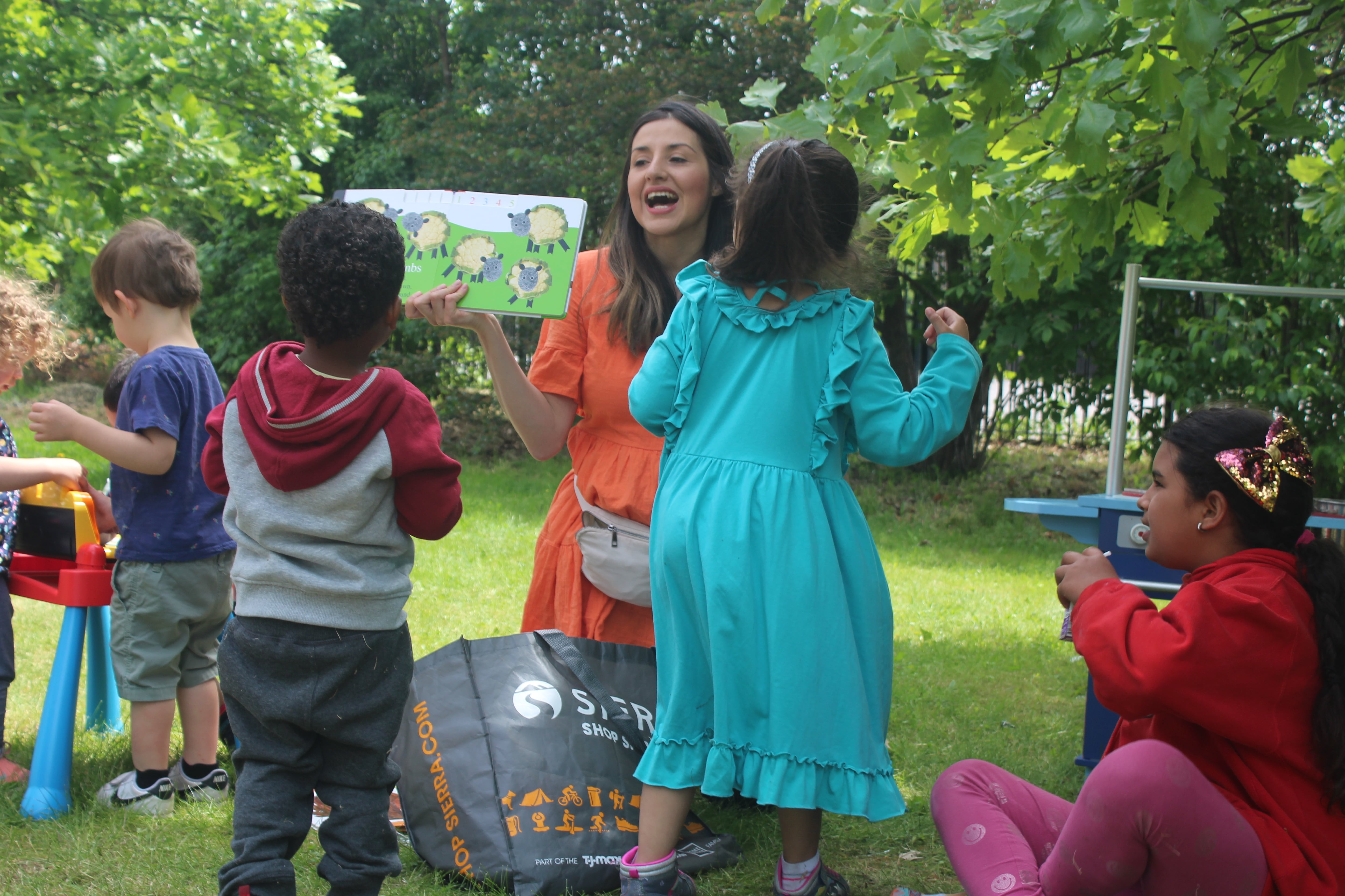 A woman with brown hair and a red dress laughs with four children of different ages while standing on grass in front of trees in a park.