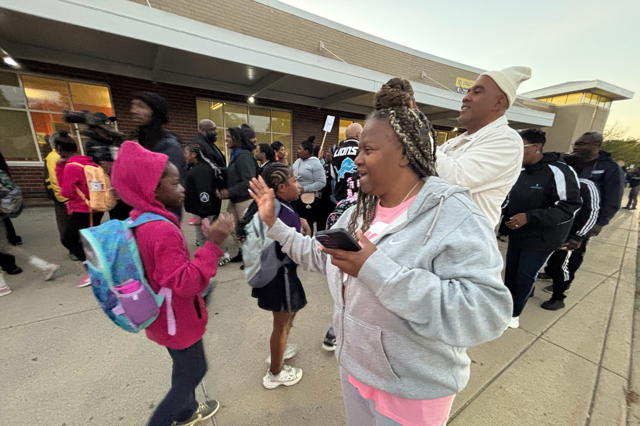 A photograph of a woman giving a high five to a student among a crowd of adults and students outside of a school building. People are wearing hoodies and sweaters.