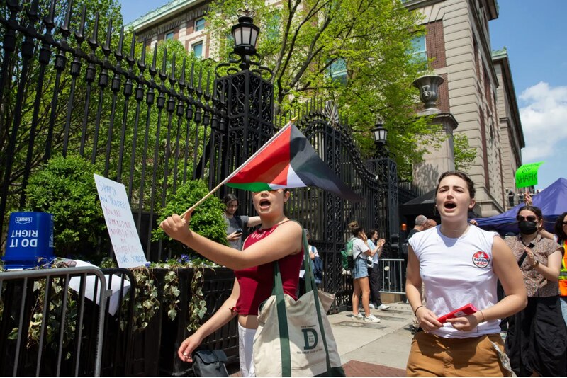 Two college students, one holding a Palestinian flag, the other chanting, walk outside an iron gate.