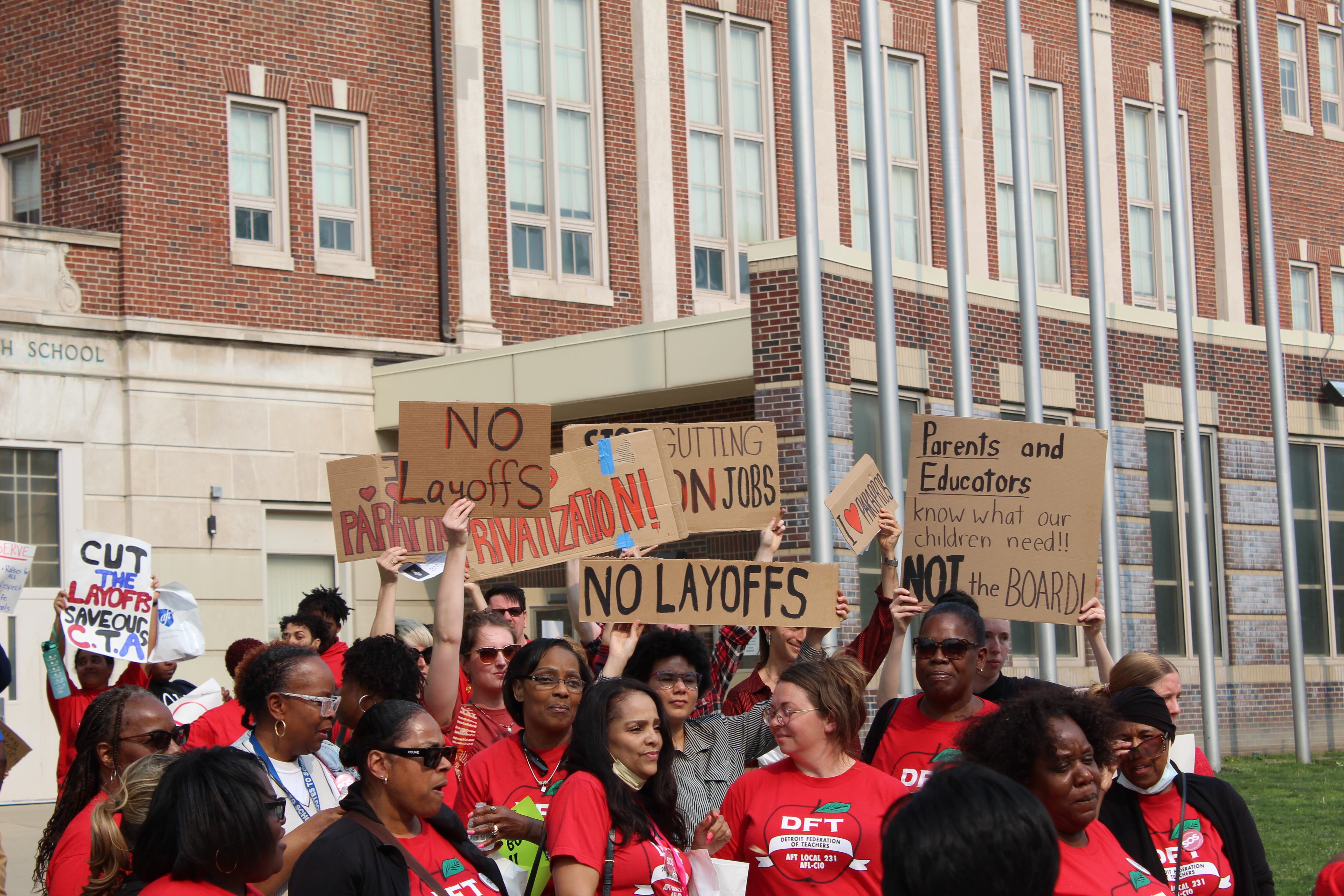 People all wearing red T-shirts are holding up signs that read “No layoffs.”