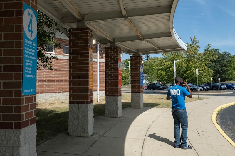A man wearing a blue shirt and jeans talks on the phone while standing outside of an entrance of a school.