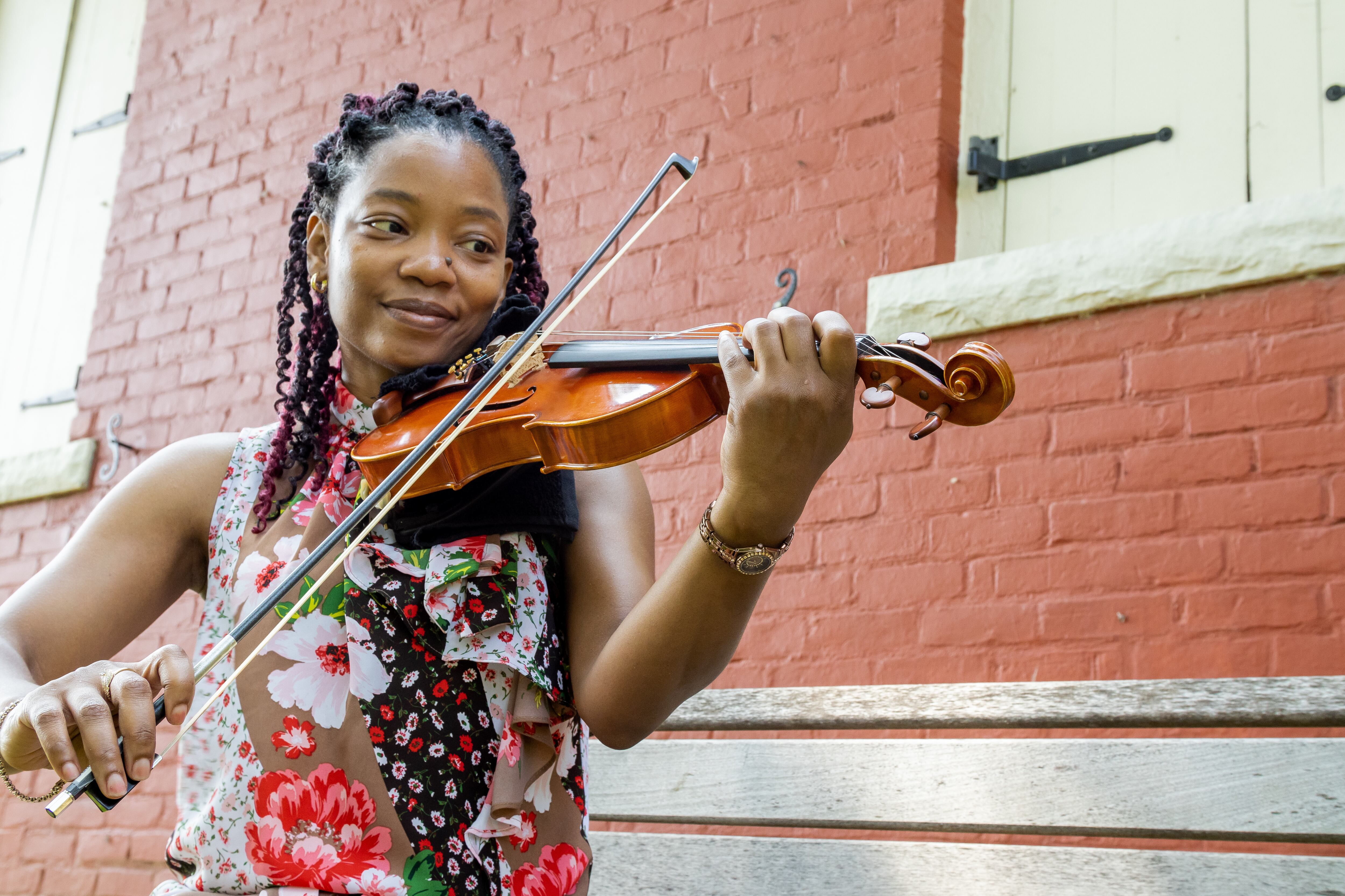 A woman stands playing a violin, with a brick building in the background.