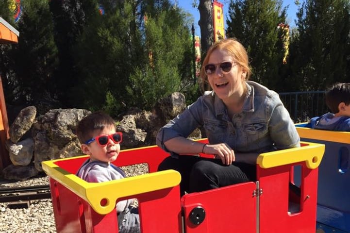 A woman wearing sunglasses sits in a small red train car outside with her son who is also wearing sunglasses.