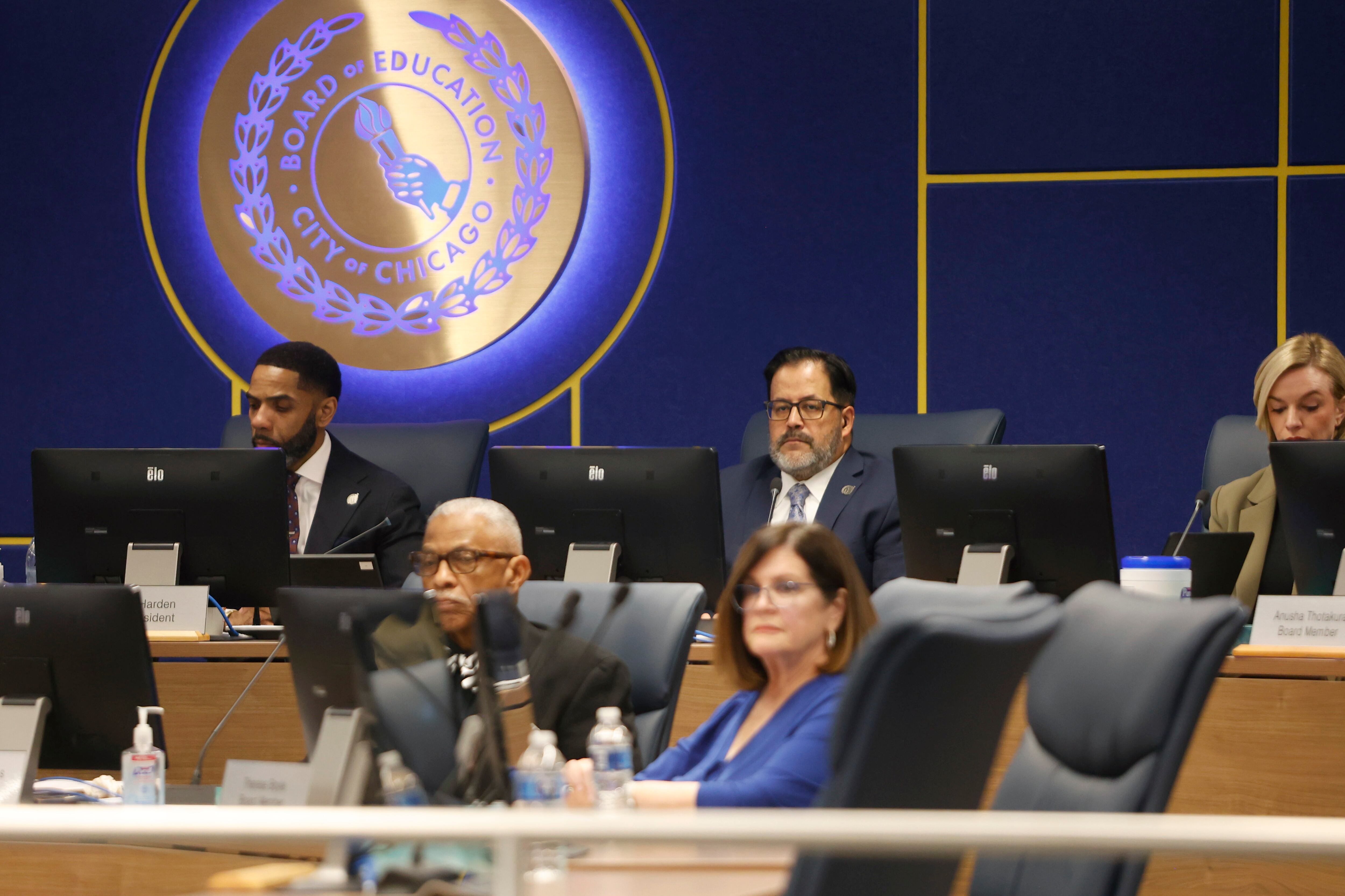 People in business clothes sit in a large conference room with a blue wall and an illuminated circular sign in the background.
