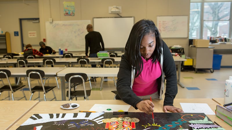 A girl wearing a black hoodie and a pink polo shirt works on an art project while other students speak with a teacher on the other side of the classroom.