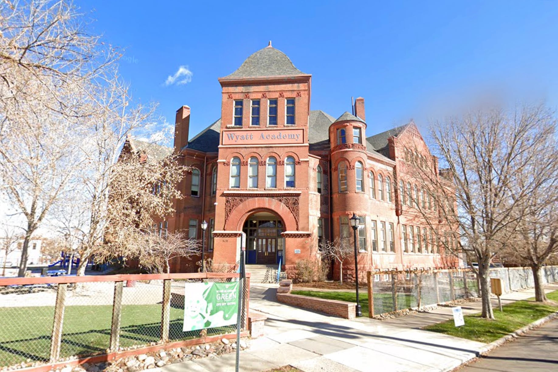 A large red stone building sits between dormant trees with a sign out front and blue skies in the background.