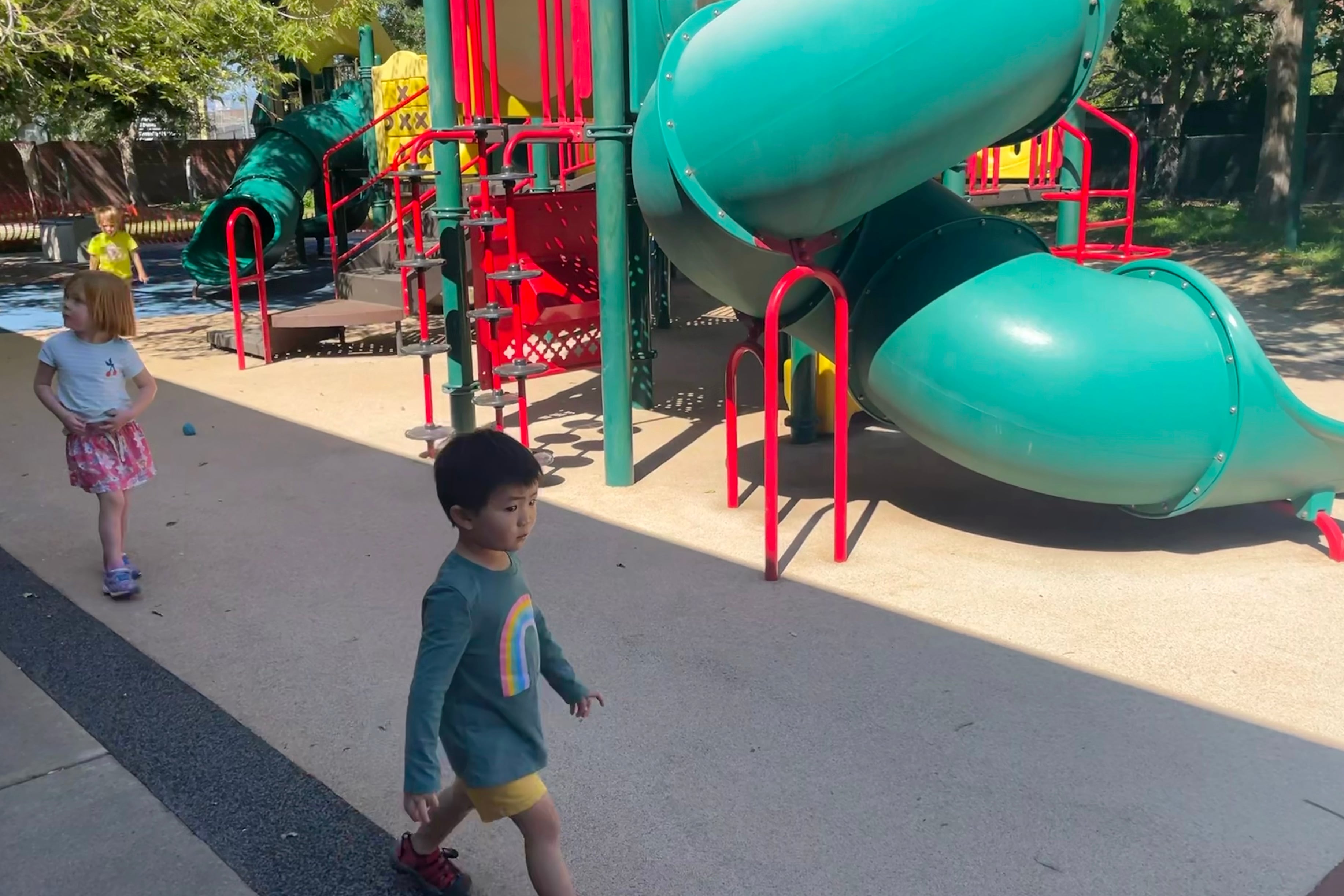 A preschool boy with a rainbow on his shirt walks past a green and red outdoor play structure.