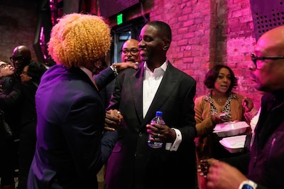 A Black man in a suit reach out to shake a hand with another man in a room full of people.
