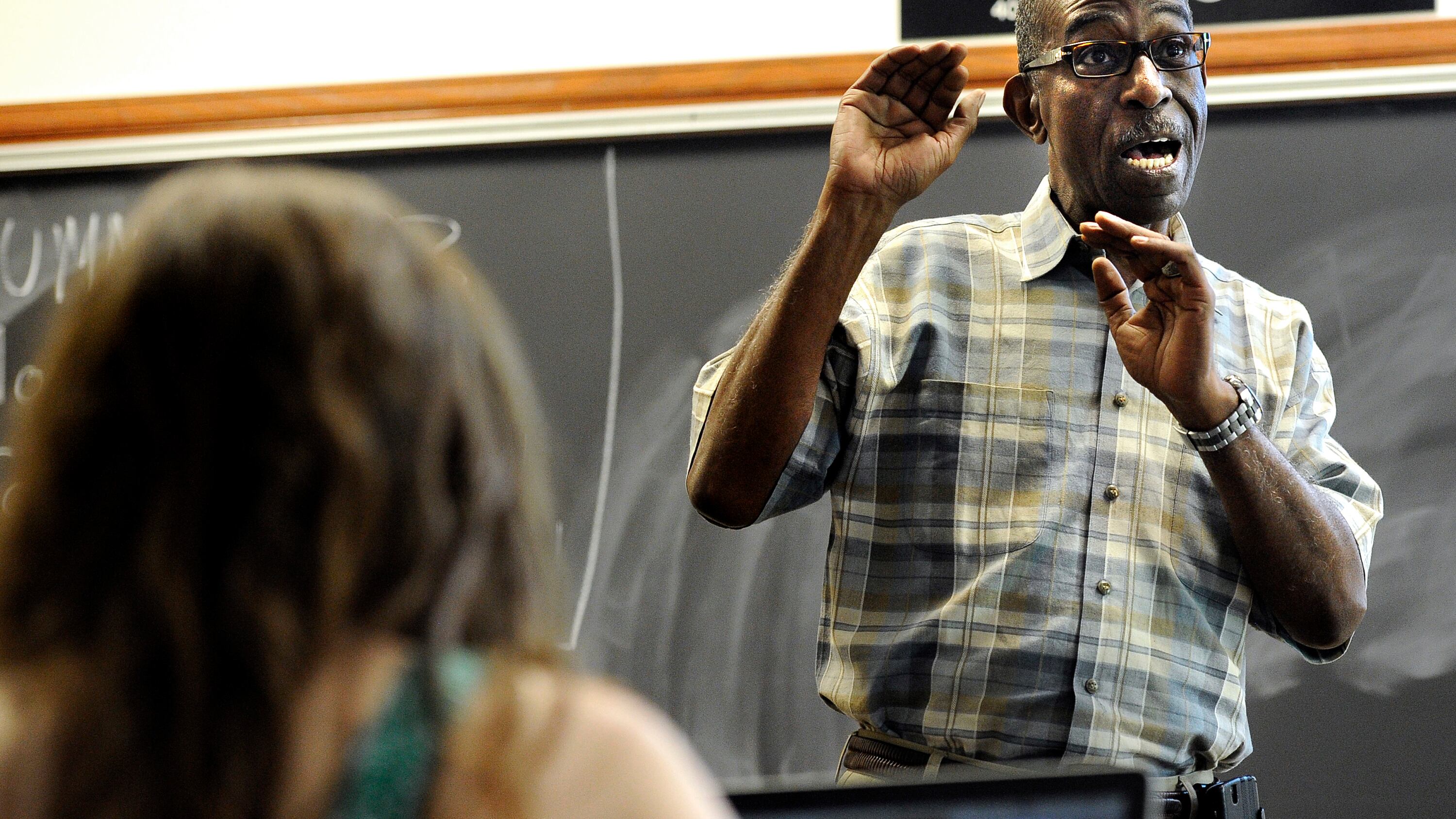 A teacher wearing a checkered shirt and glasses emphatically lectures as a student listens in the foreground.