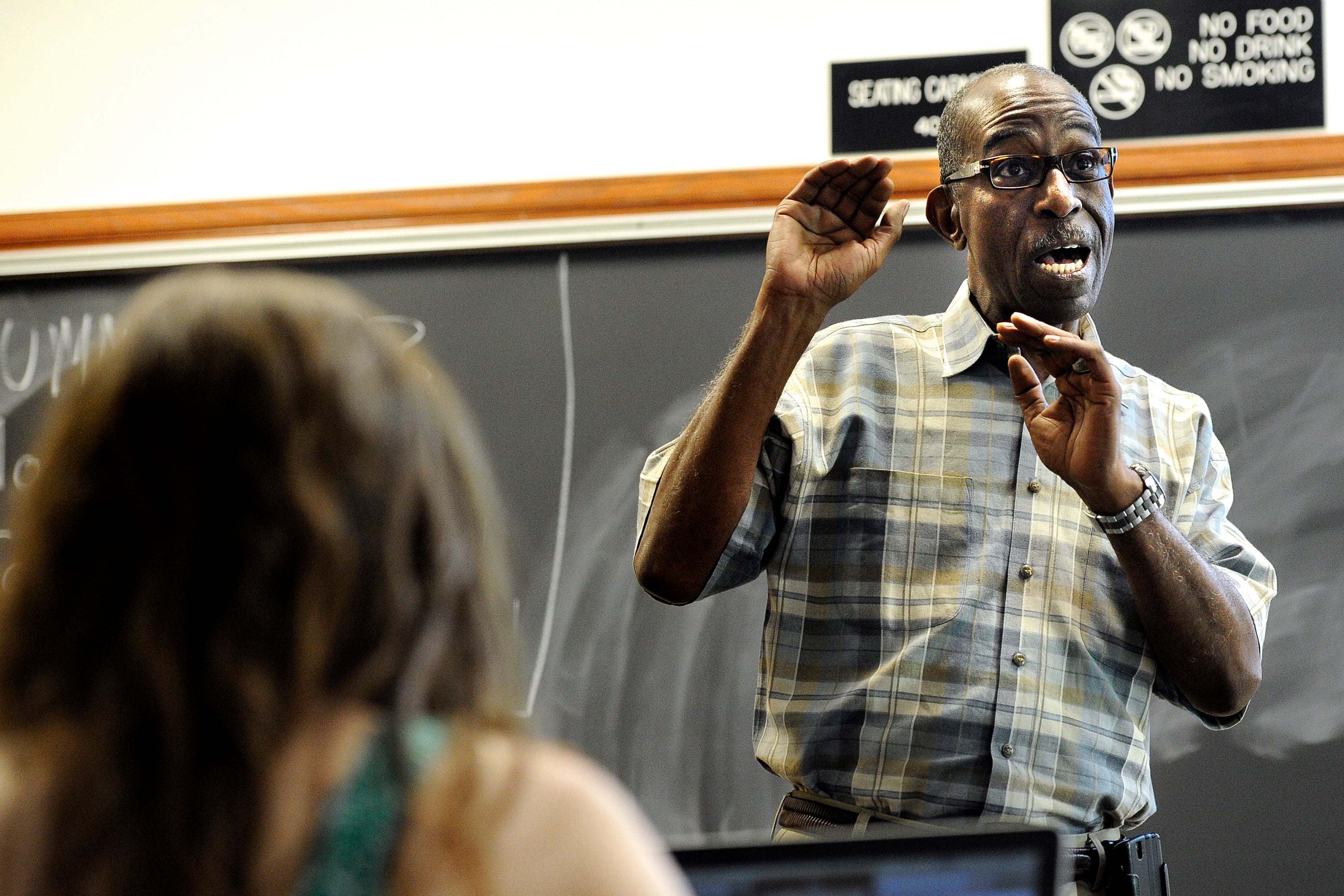 A teacher wearing a checkered shirt and glasses emphatically lectures as a student listens in the foreground.