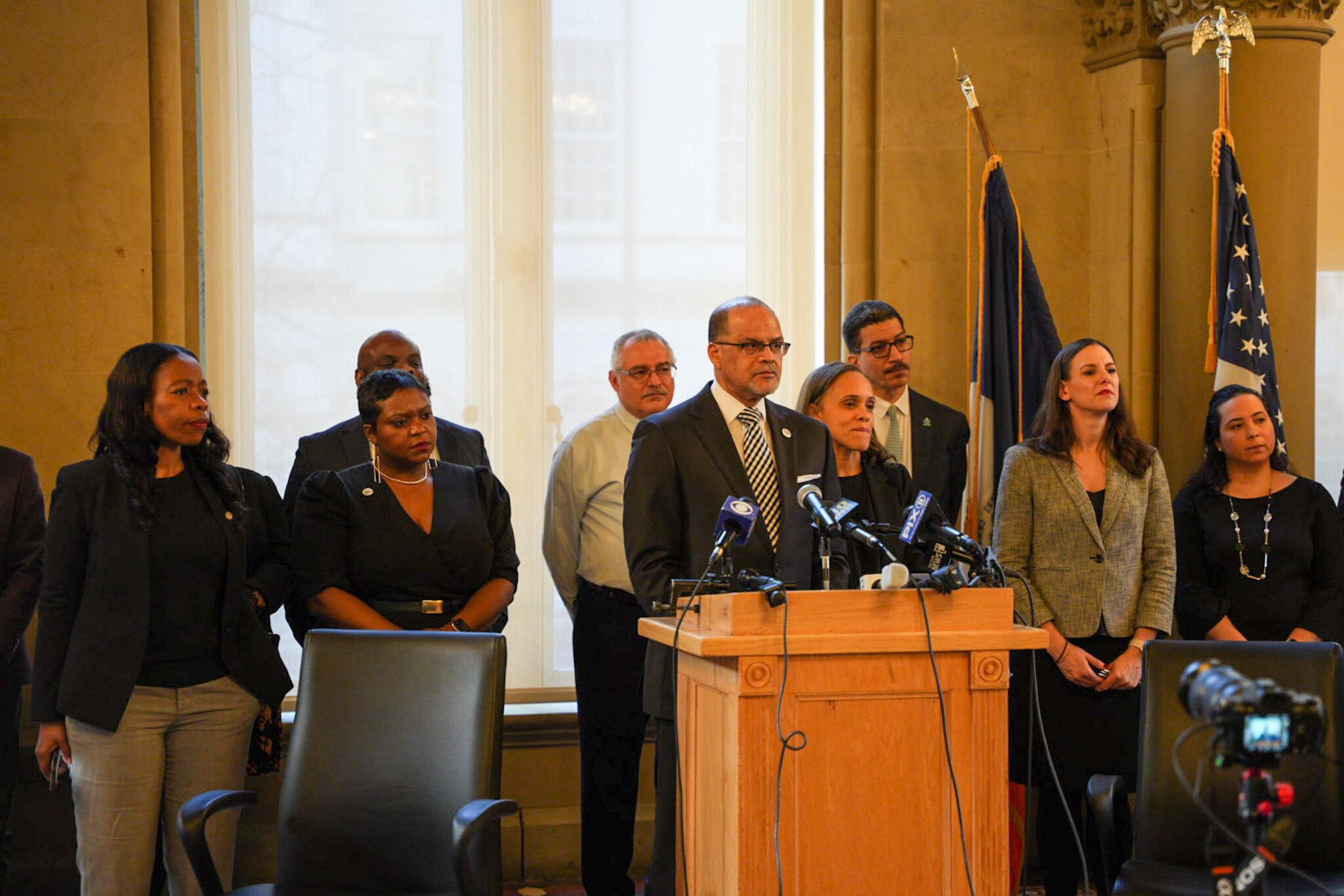 A man stands at a podium at a press conference surrounded by a line of people behind him.