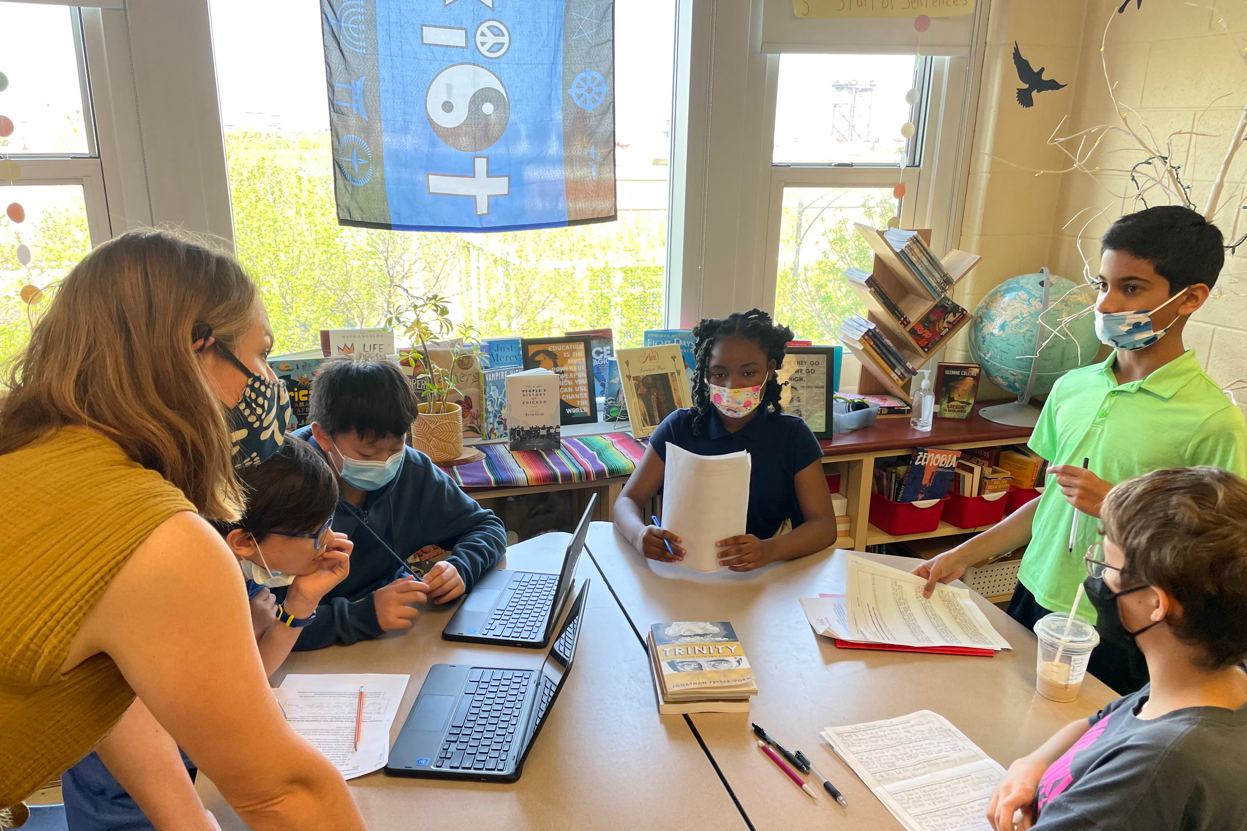 A woman wearing a face mask talks with children wearing face masks and sitting around a round table with laptops and paperwork.