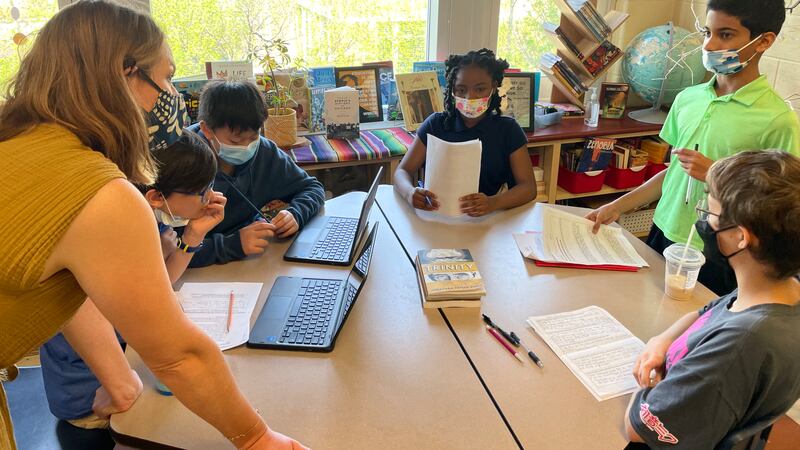 A woman wearing a face mask talks with children wearing face masks and sitting around a round table with laptops and paperwork.
