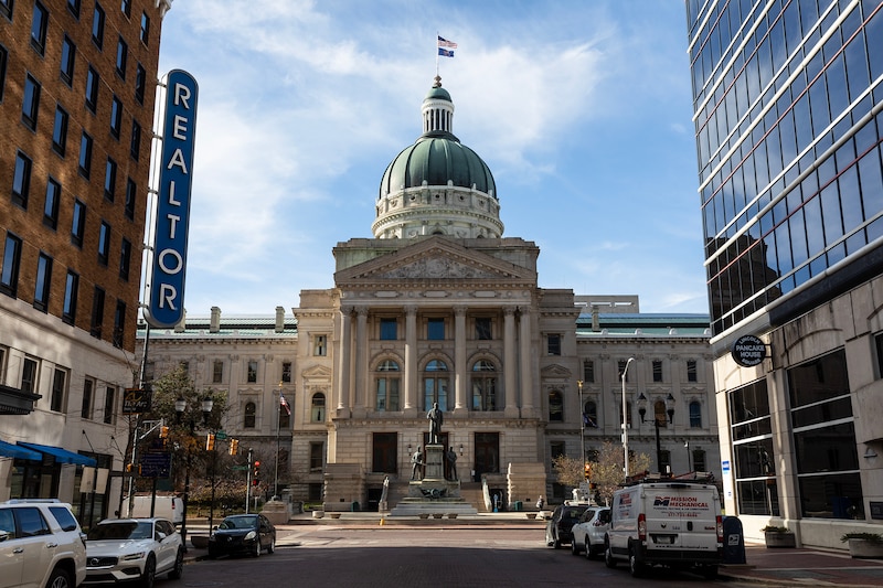 A large tan stone building with a green dome roof is in the background with tall buildings in the foreground with cars in the street.