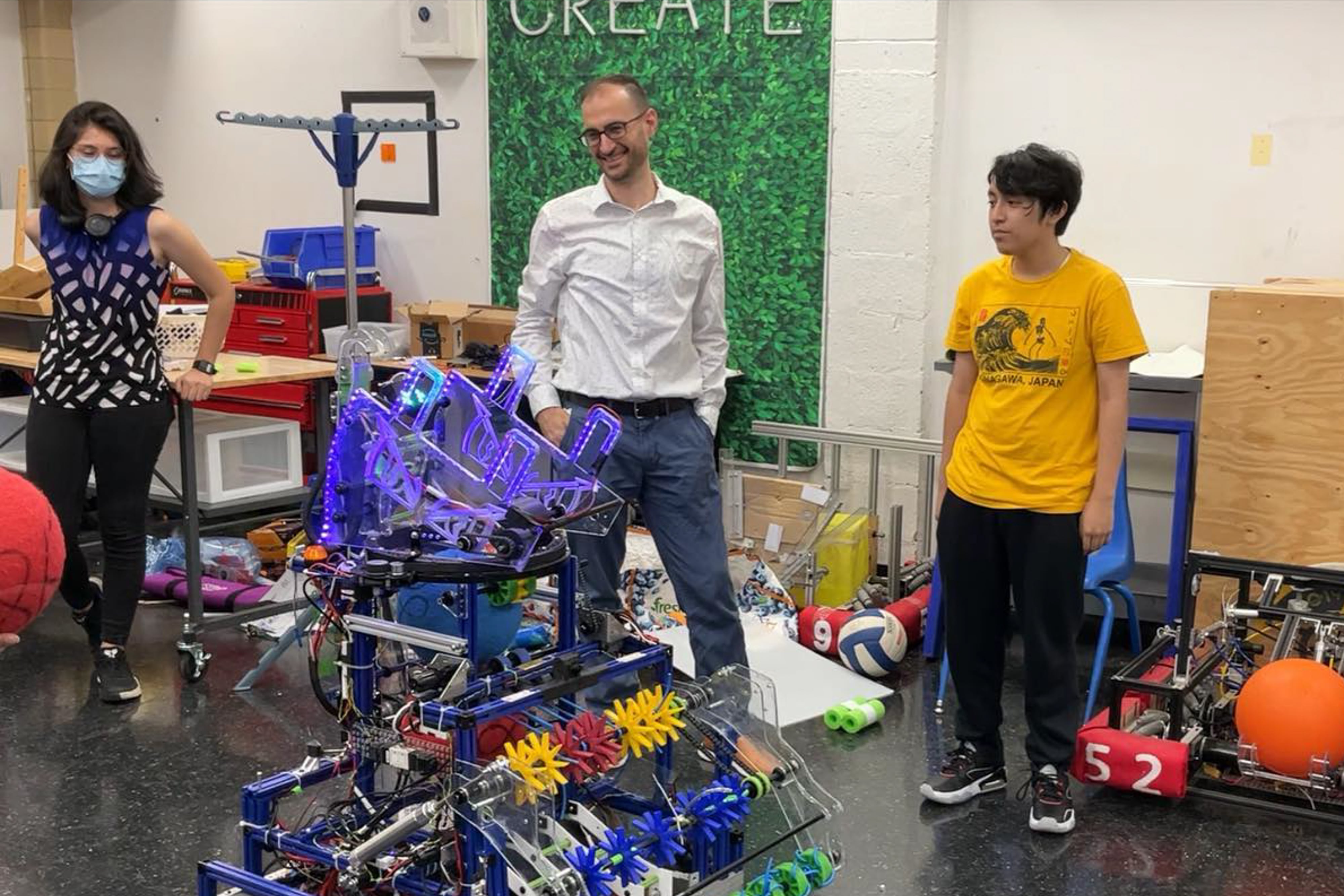 A woman wearing a face mask, a man in a white button-down shirt and a teen in a yellow T-shirt stand in a classroom with a school-made robot.
