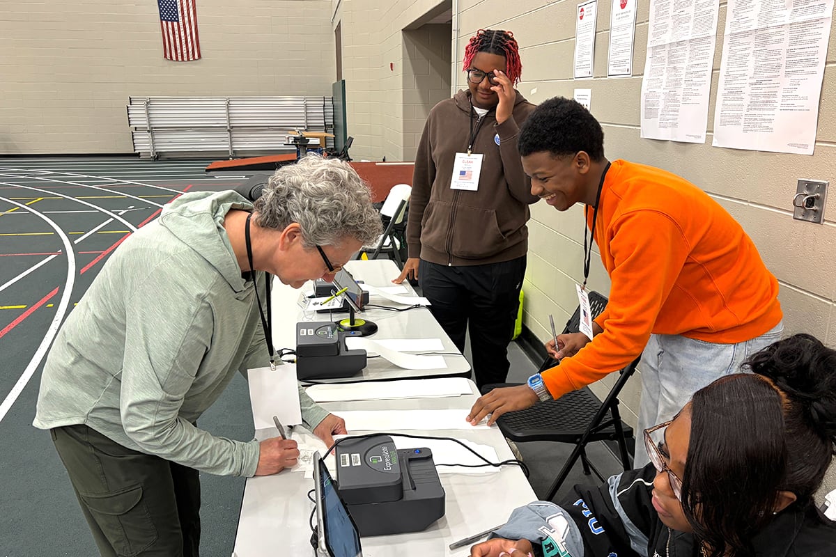 A photograph of an adult signing in at polling location while high school students are working as election workers at a gym.