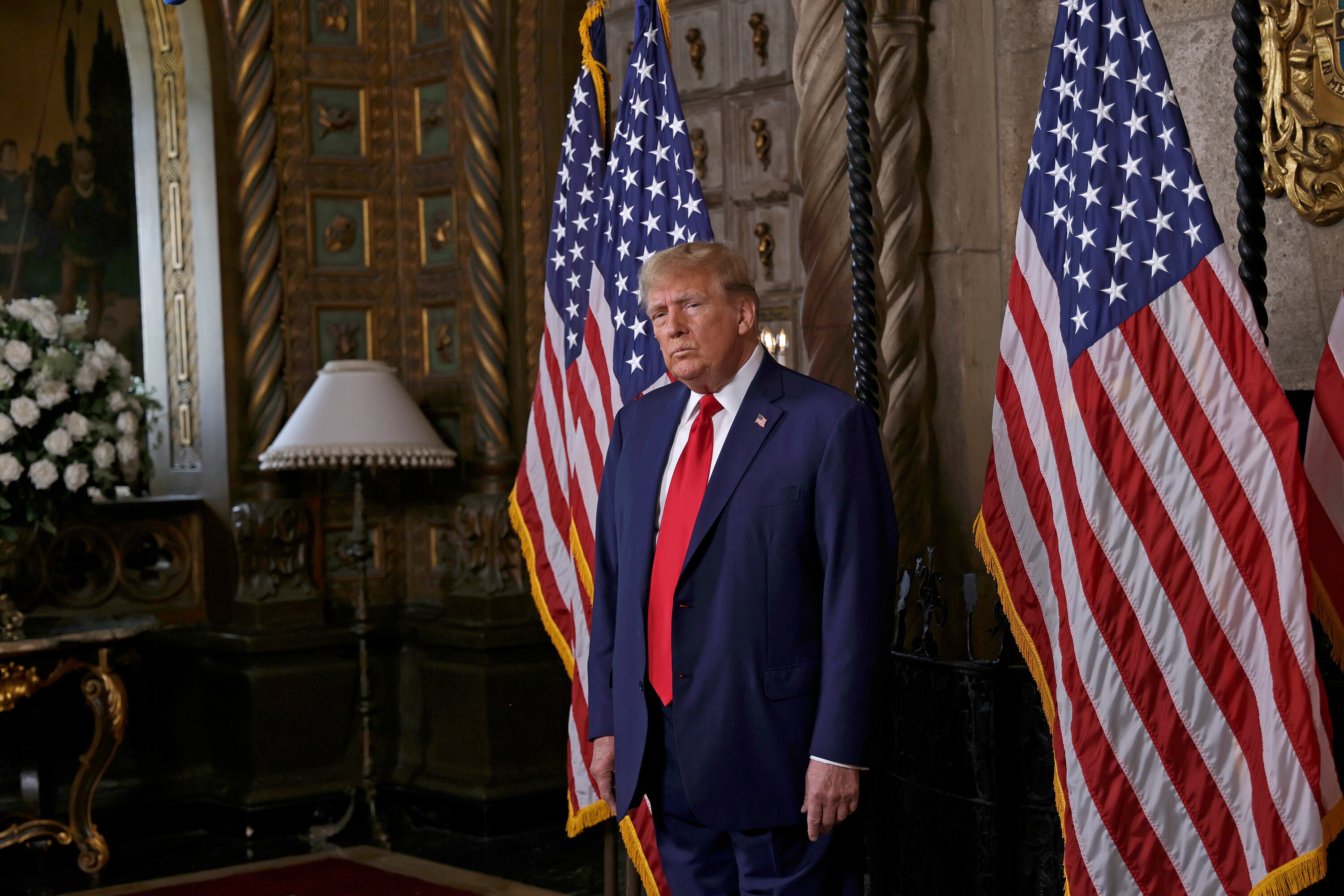 Donald Trump wearing a dark suit and red tie stands between two American flags with a lamp and flowers to the left of him and wooden doors in the background.