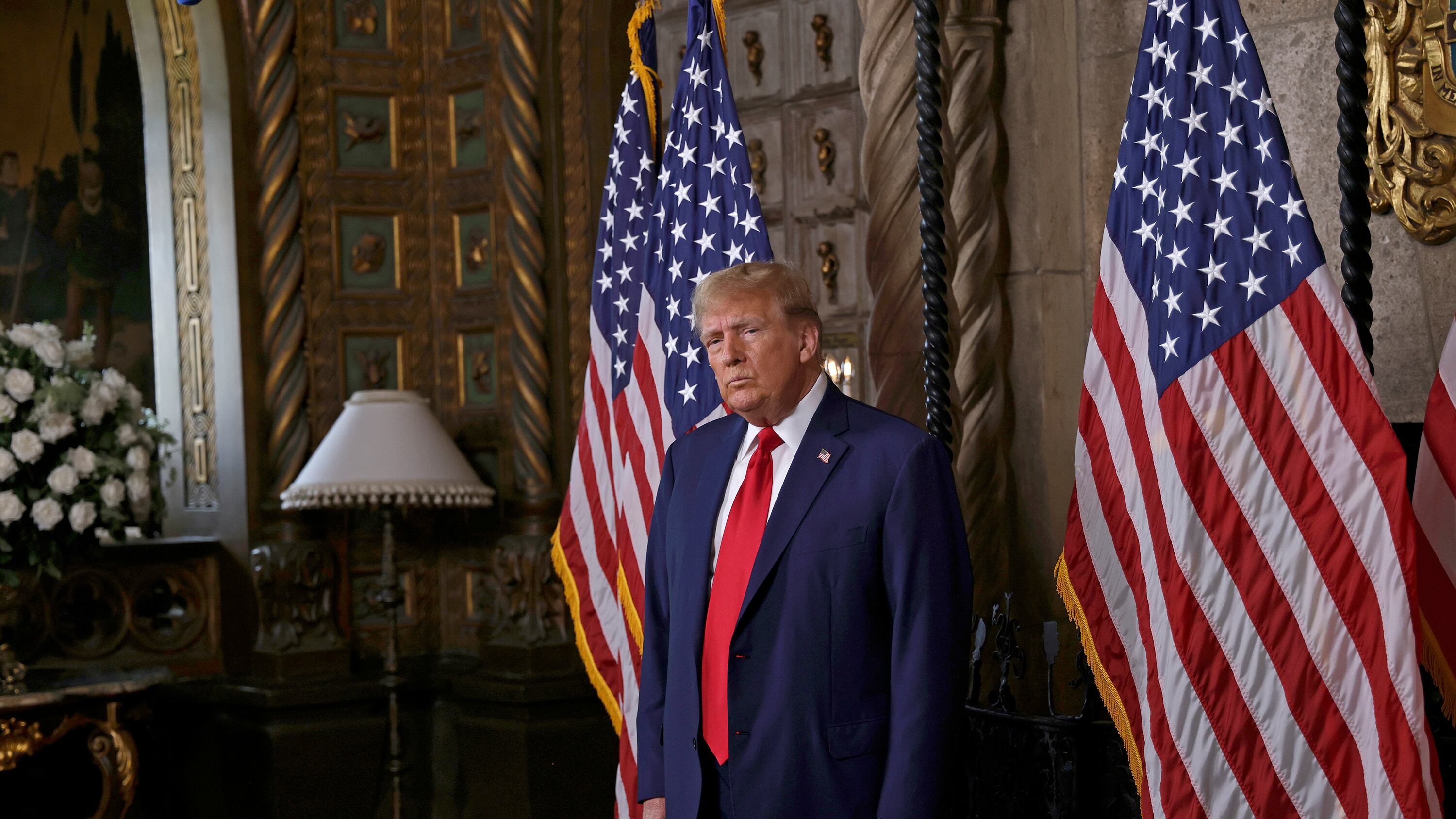 Donald Trump wearing a dark suit and red tie stands between two American flags with a lamp and flowers to the left of him and wooden doors in the background.