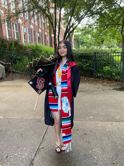 A high school senior with long dark hair and wearing a cap and gown poses for a portrait outside.