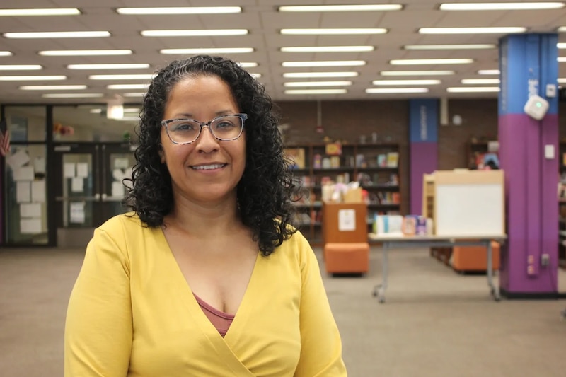 A woman with glasses and wearing a yellow top stands in a room with bookshelves