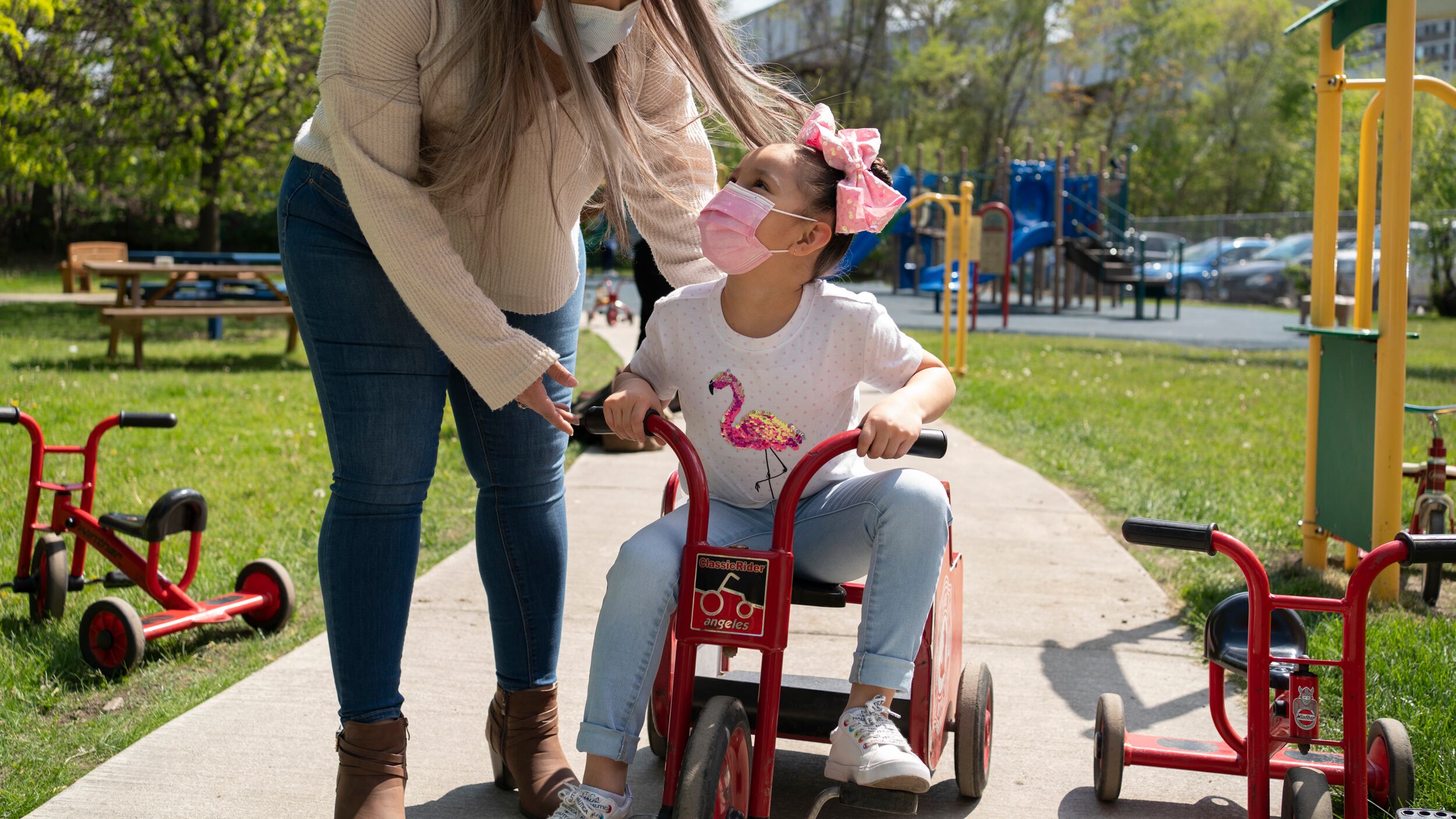 A mother wearing jeans, tan shirt and protective mask pushes her daughter on a red tricycle. The daughter is wearing a white flamingo shirt, pink mask, pink bow and jeans.