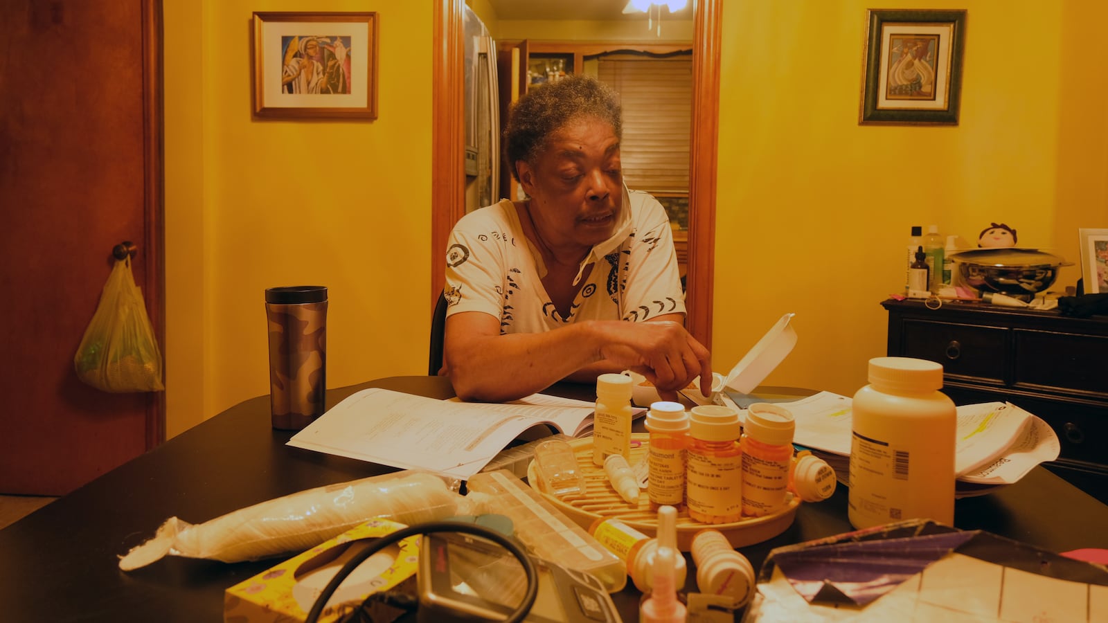 A woman sits at her kitchen table with several prescription pill bottles and papers on the table.