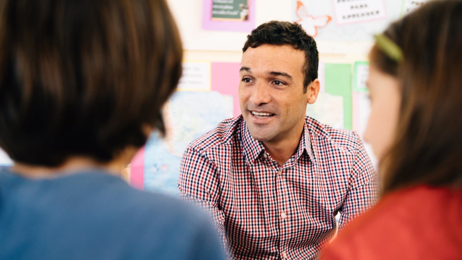 Man in button down shirt talks to young children in a school setting. 