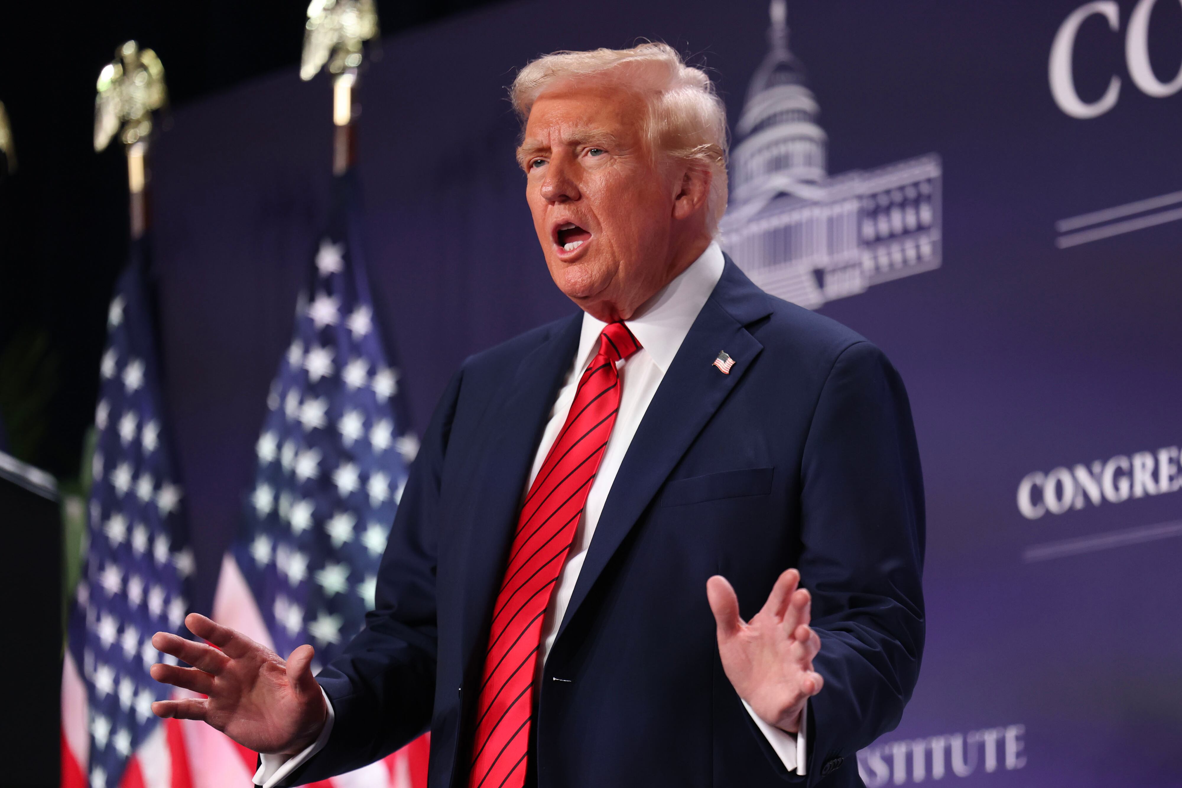 President Donald Trump wearing a dark blue suit and a red tie speaks in front of a dark background with white words and drawings with two American flags in the background.