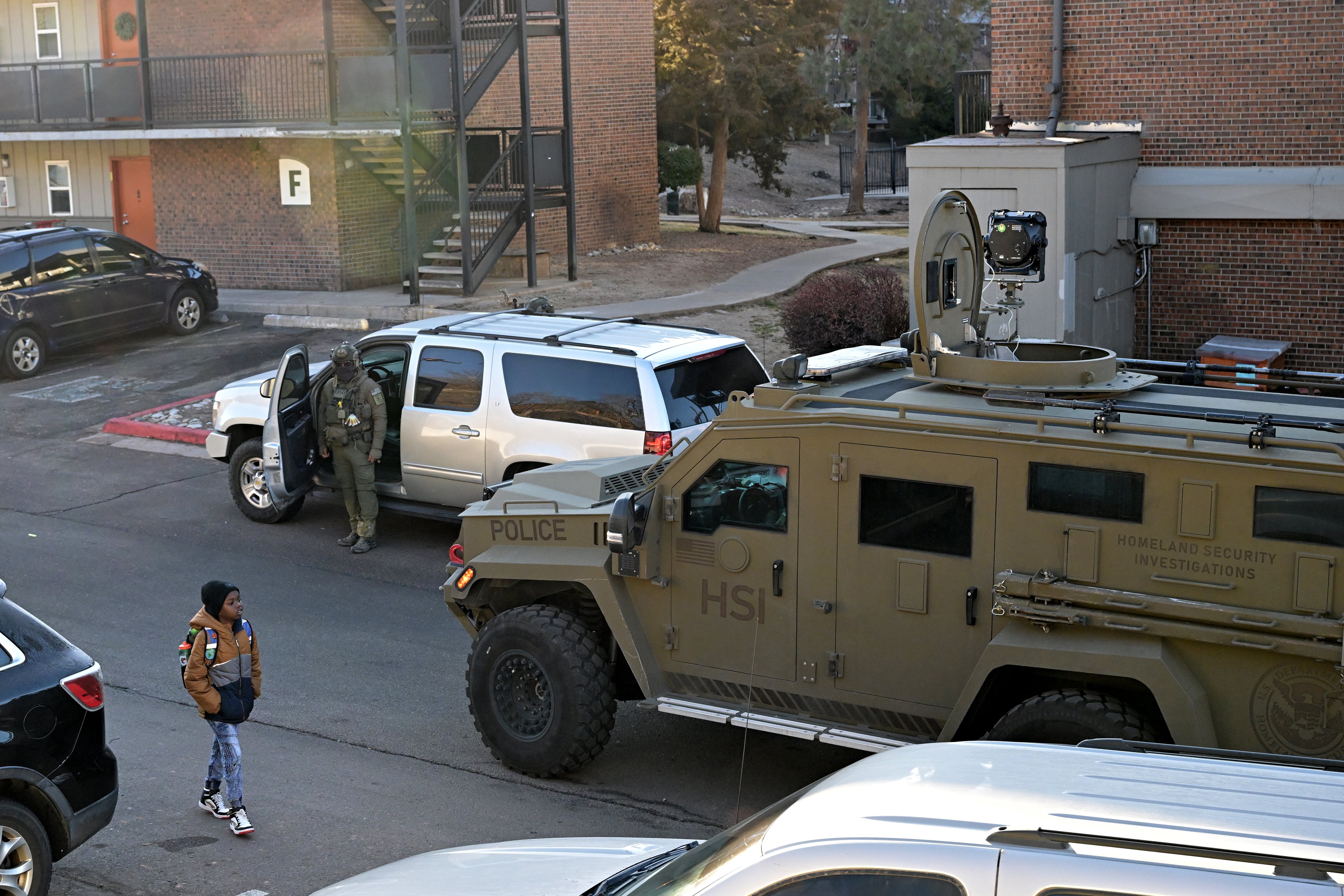 A young boy carrying a backpack and dressed in a coat and hat walks past a man in tactical gear next to an armored vehicle and an SUV in an apartment complex.