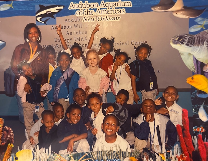 A photograph of a Black teacher posing next to a group of young students all posing for a photograph in front of an aquarium poster.