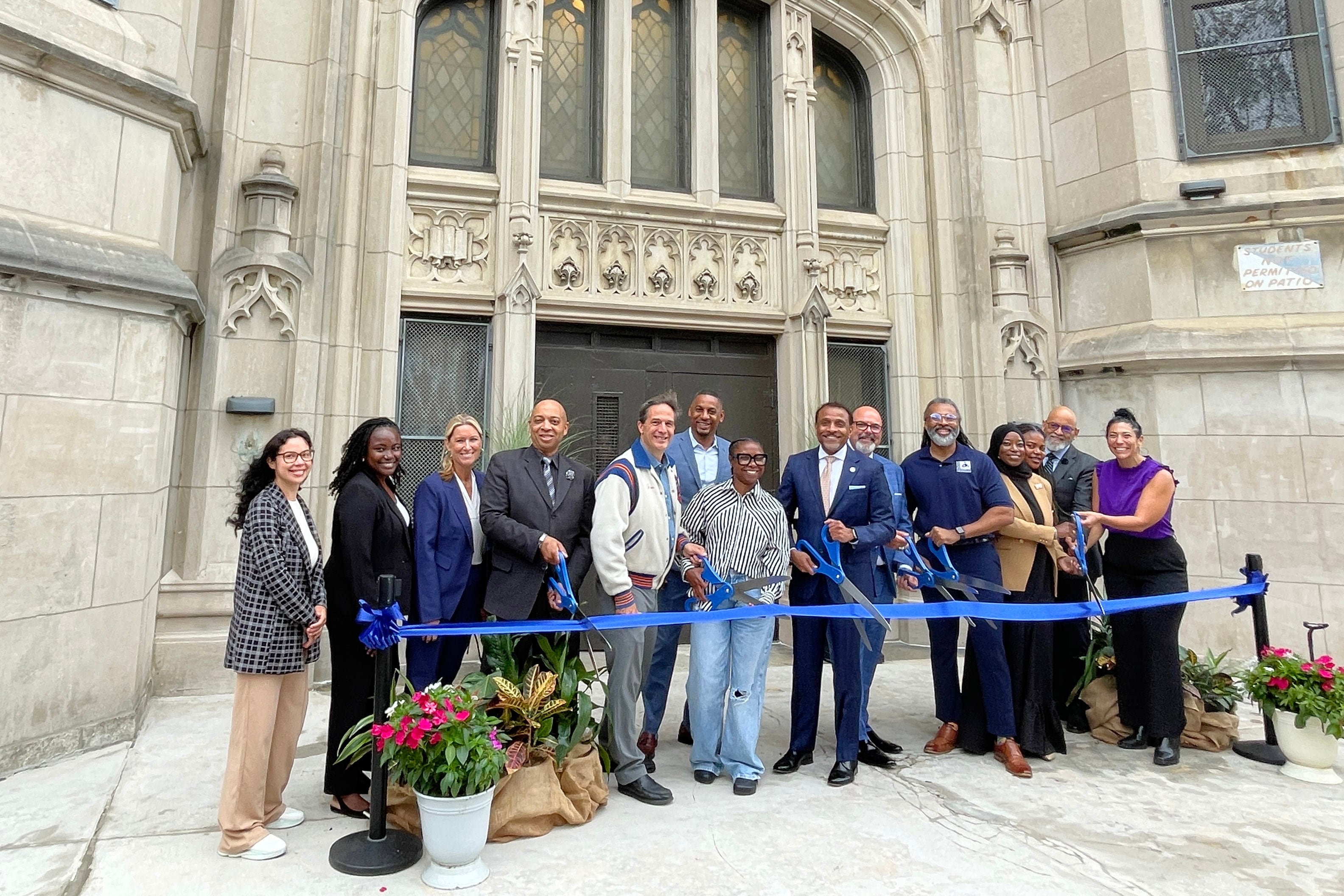 A photograph of a line of people standing behind a blue ribbon and in front of a large stone school building.