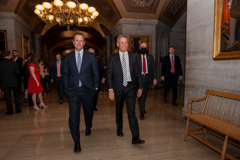 Two men wearing suits walk in a crowded hallway with a chandalier in the backdrop.