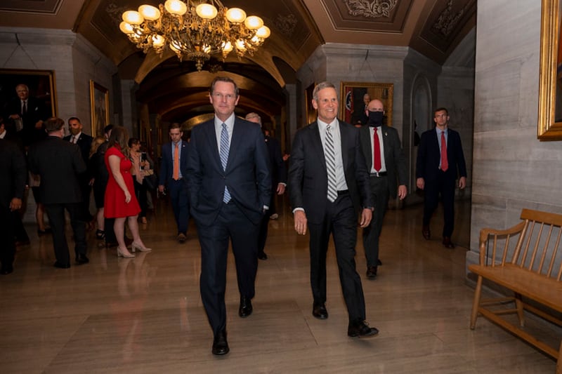 Two men wearing suits walk in a crowded hallway with a chandalier in the backdrop.