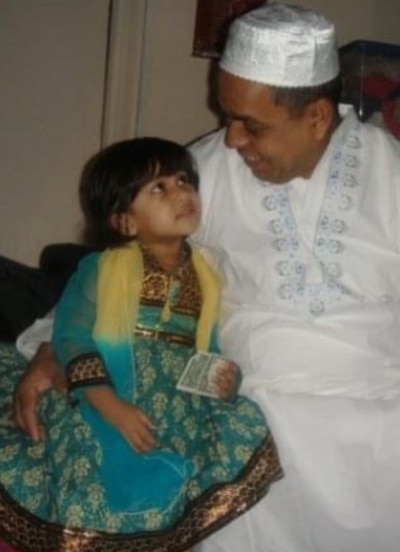 Photo of a father wearing a white top and a white hat sits with his young daughter, who is wearing a multicolored dress.