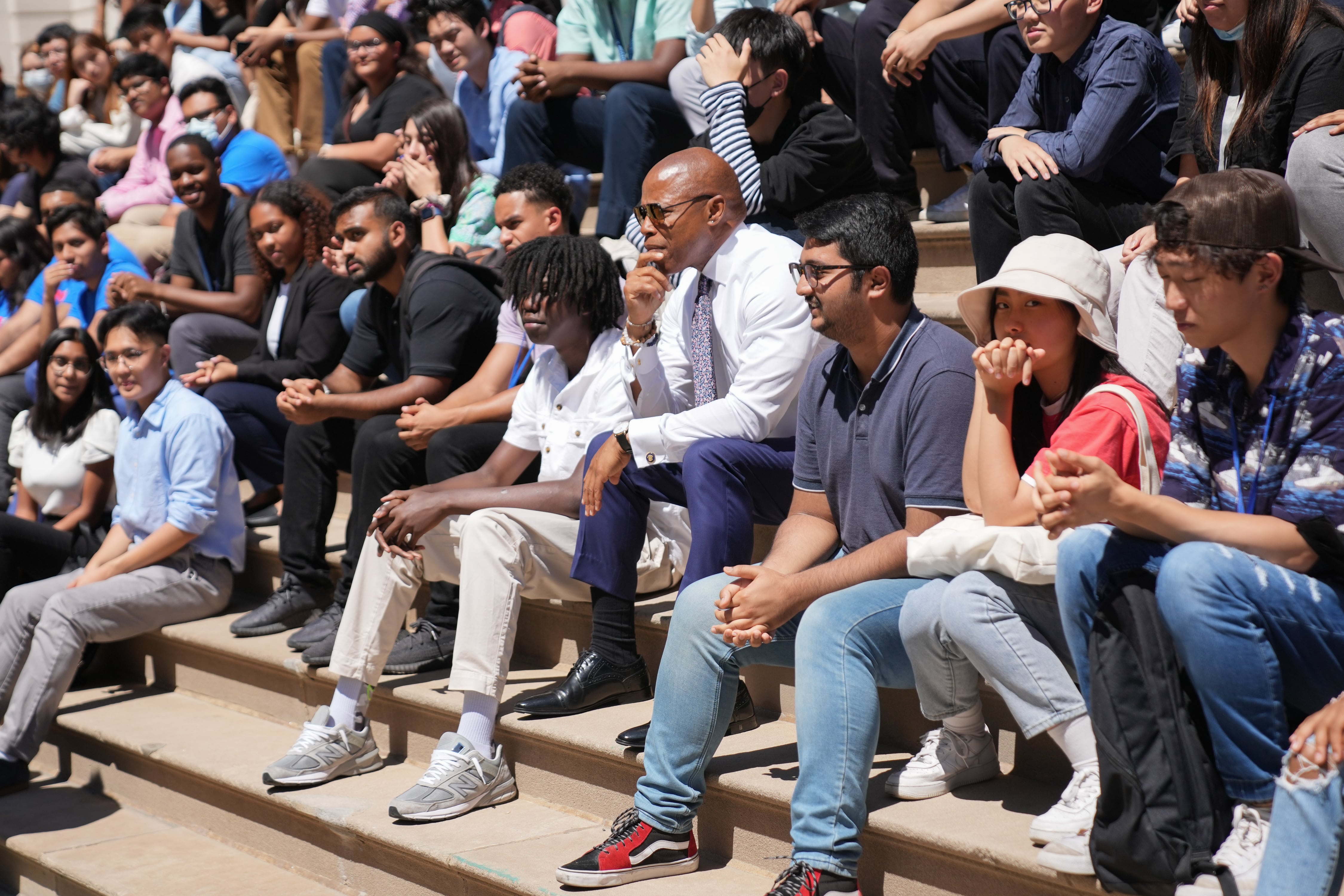 A man in a shirt and tie sits on steps flanked by a bunch of teens.