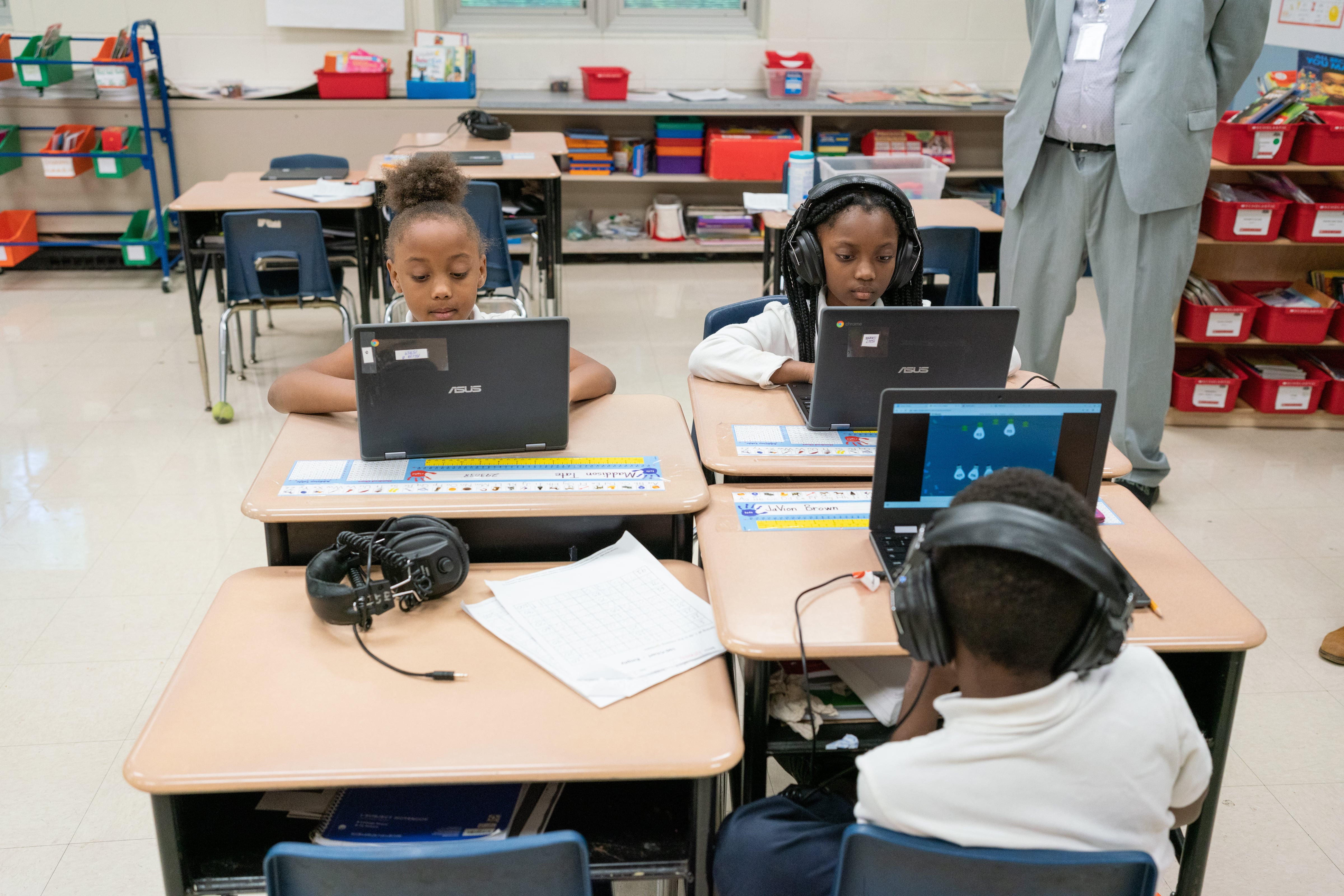 Three young students sit together at four desks and work on computers while a teacher stands behind to supervise them. One desk is empty.