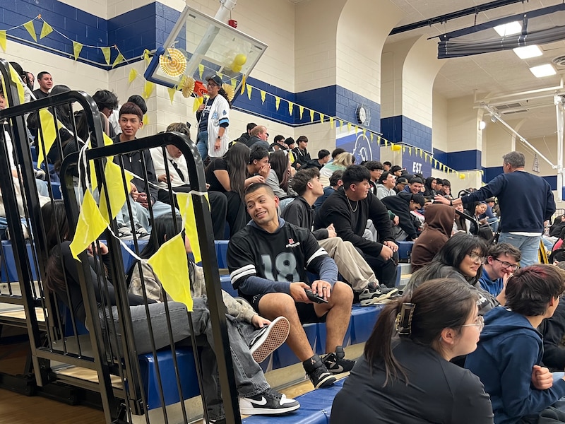 A photograph of a high school gymnasium full of high school students sitting in the bleachers.