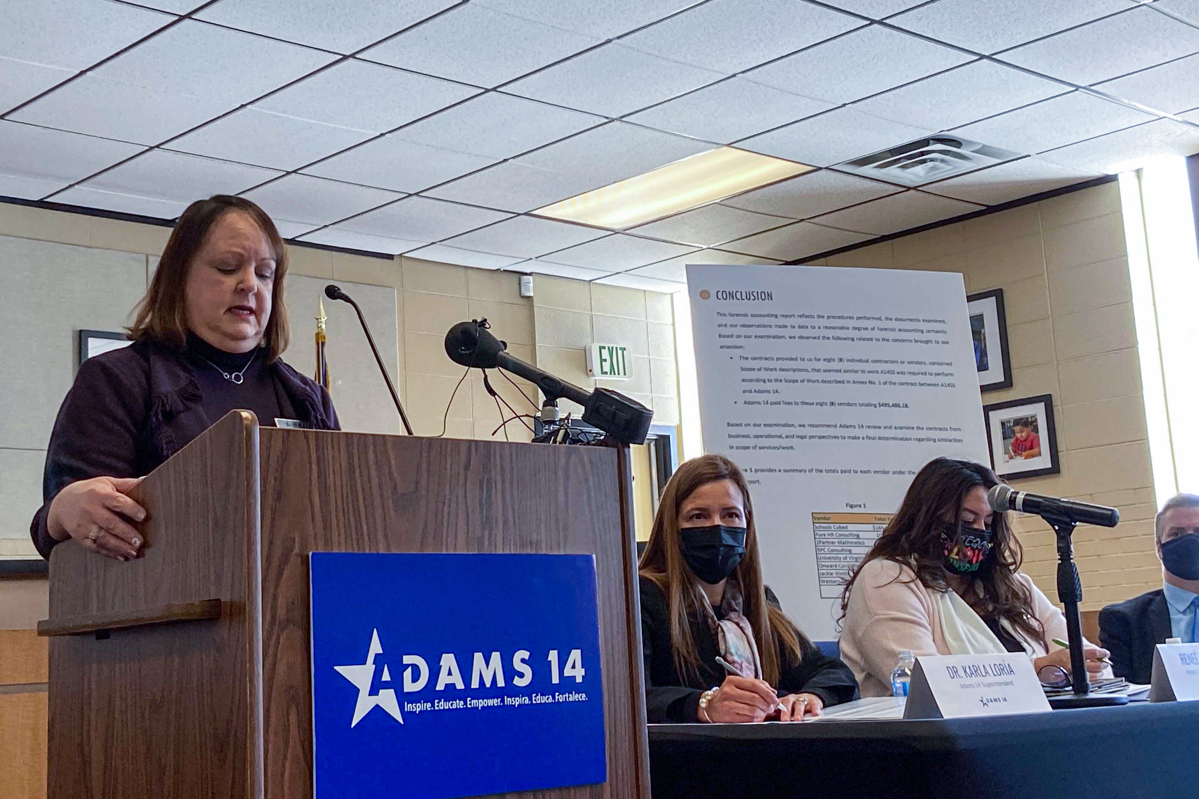 A middle-aged woman with short dark hair speaks at a podium. To her left, two other masked women, seated, look on. A blue Adams 14 school district logo is on the front of the podium.